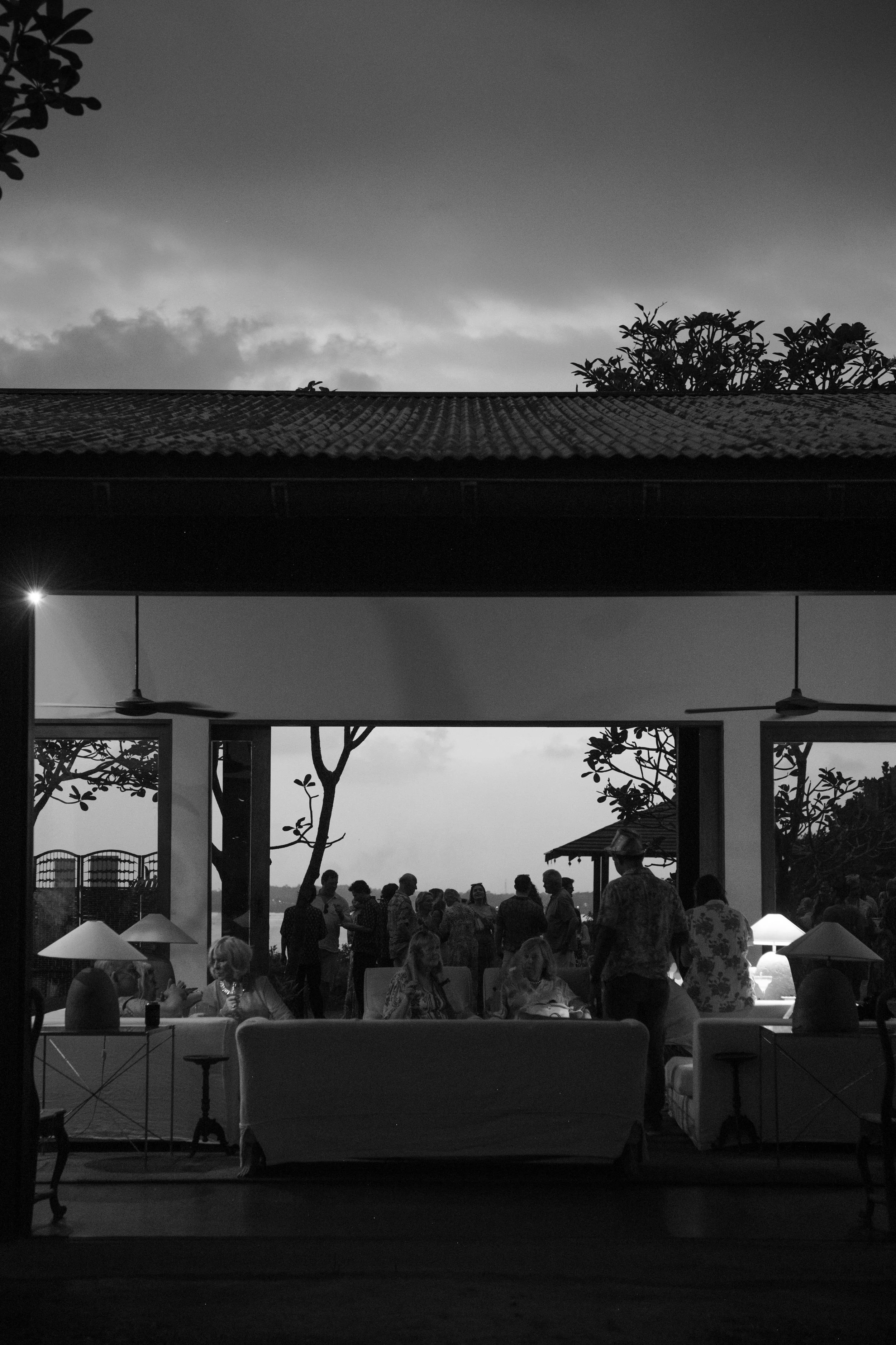People gather in a covered outdoor space, with some seated on sofas and others standing, silhouetted against the evening sky, with trees and distant horizon visible outside.