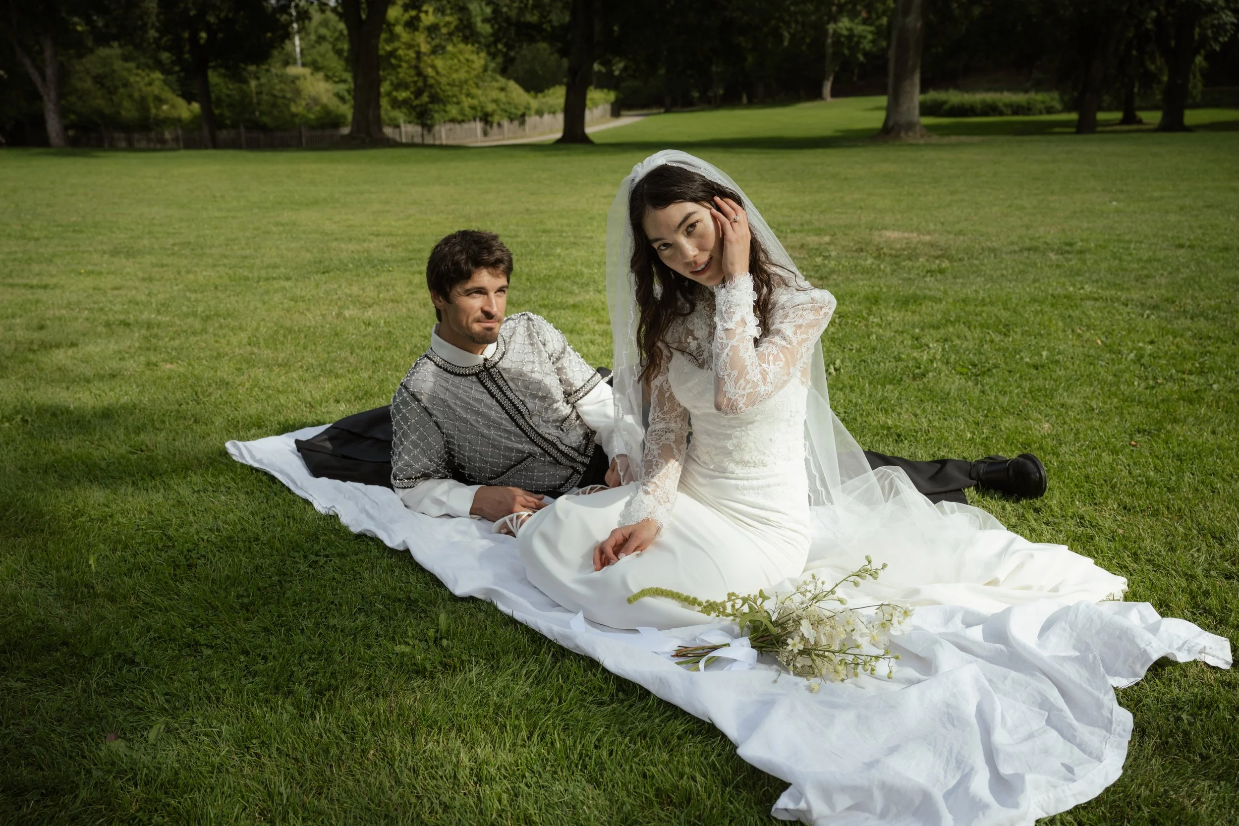 Bride in a white lace wedding gown and veil sitting on a white cloth on the grass, with groom in traditional black and white attire reclining nearby in a park with green trees.