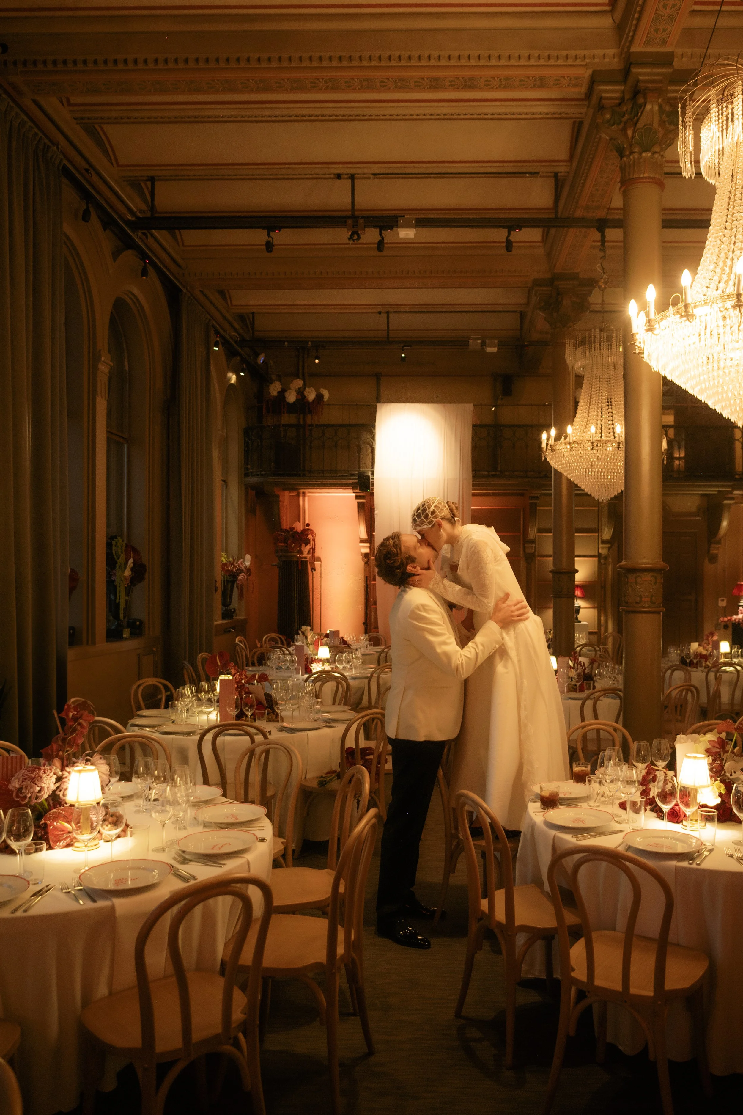 A bride and groom sharing a kiss in a decorated wedding reception hall with round tables, floral centerpieces, and chandeliers.