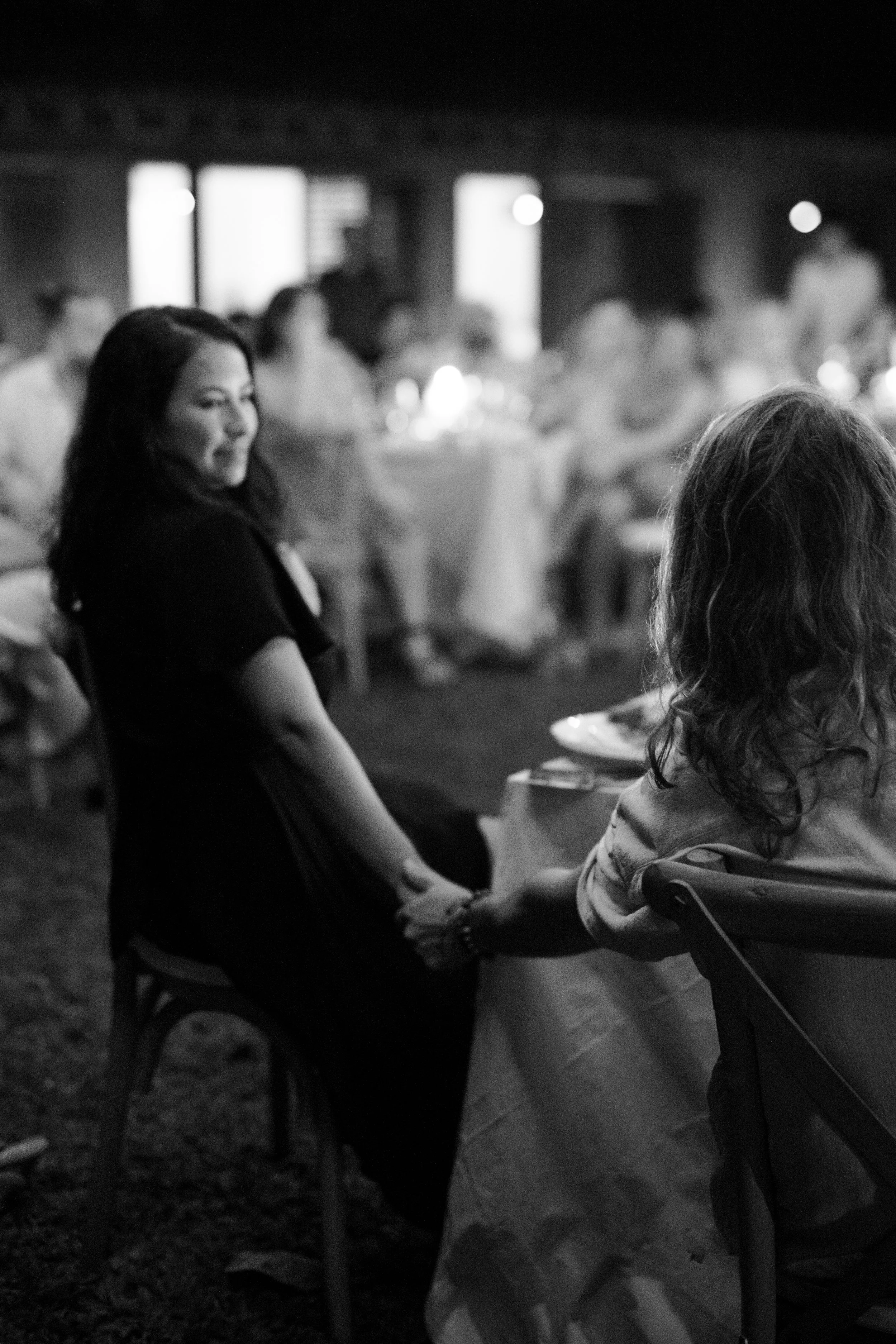 A black-and-white photo of two women holding hands at a social gathering or event, sitting at a table with other people in the background.
