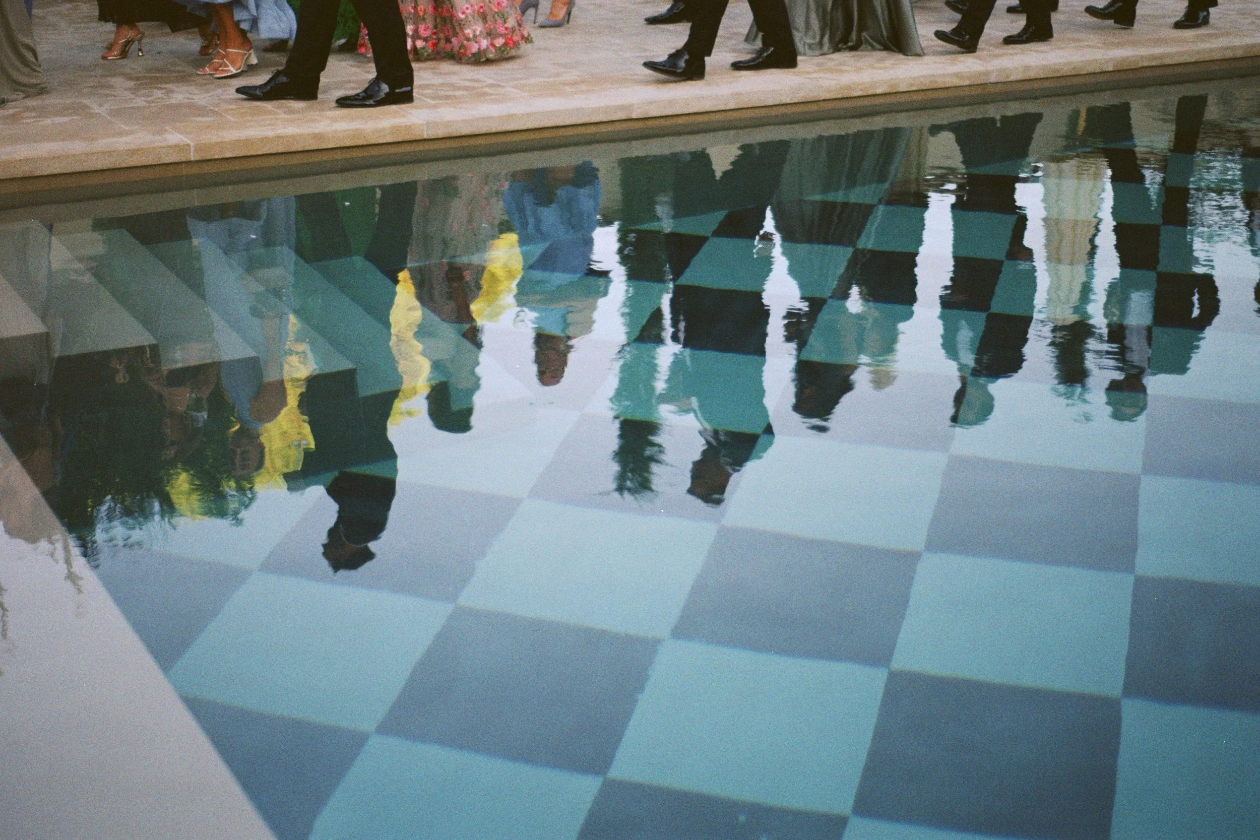 Reflection of people walking by a tiled swimming pool with checkered blue and white tiles.
