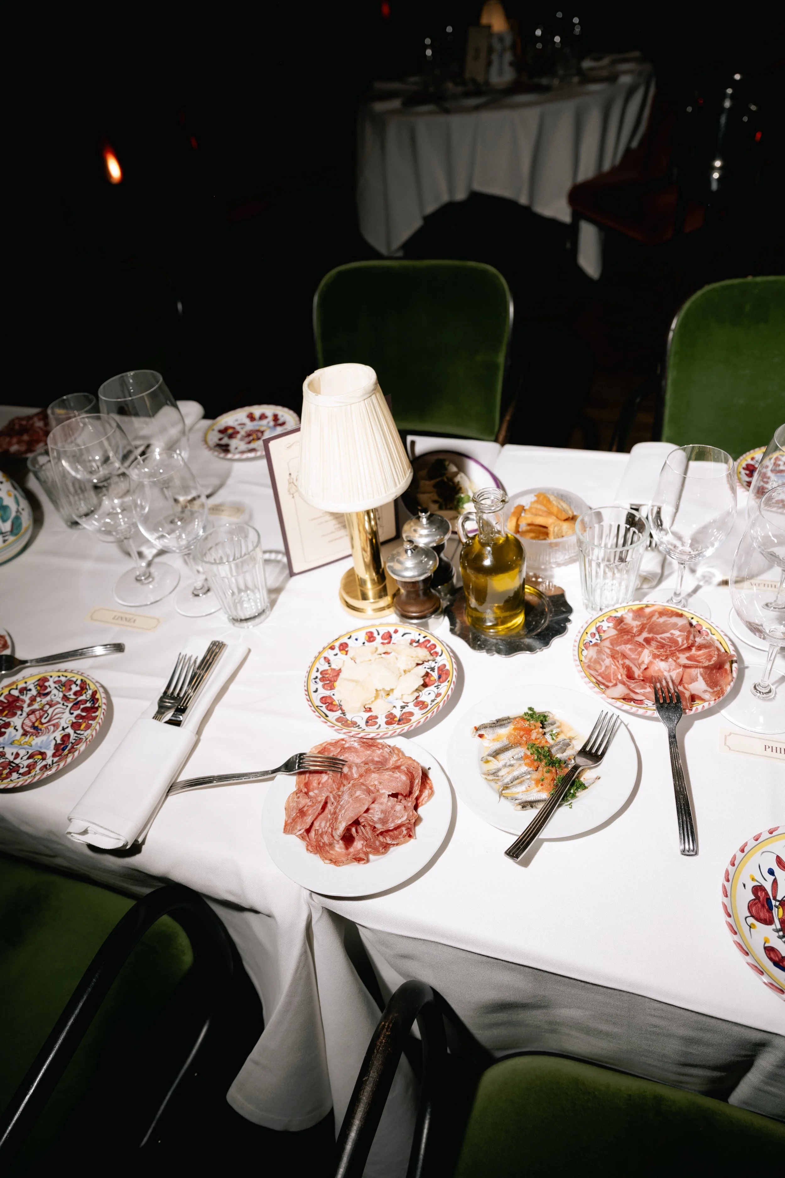 A formal dinner table set with plates of sliced meats, seafood, wine glasses, cutlery, a lamp, and condiments, in an elegant restaurant setting.
