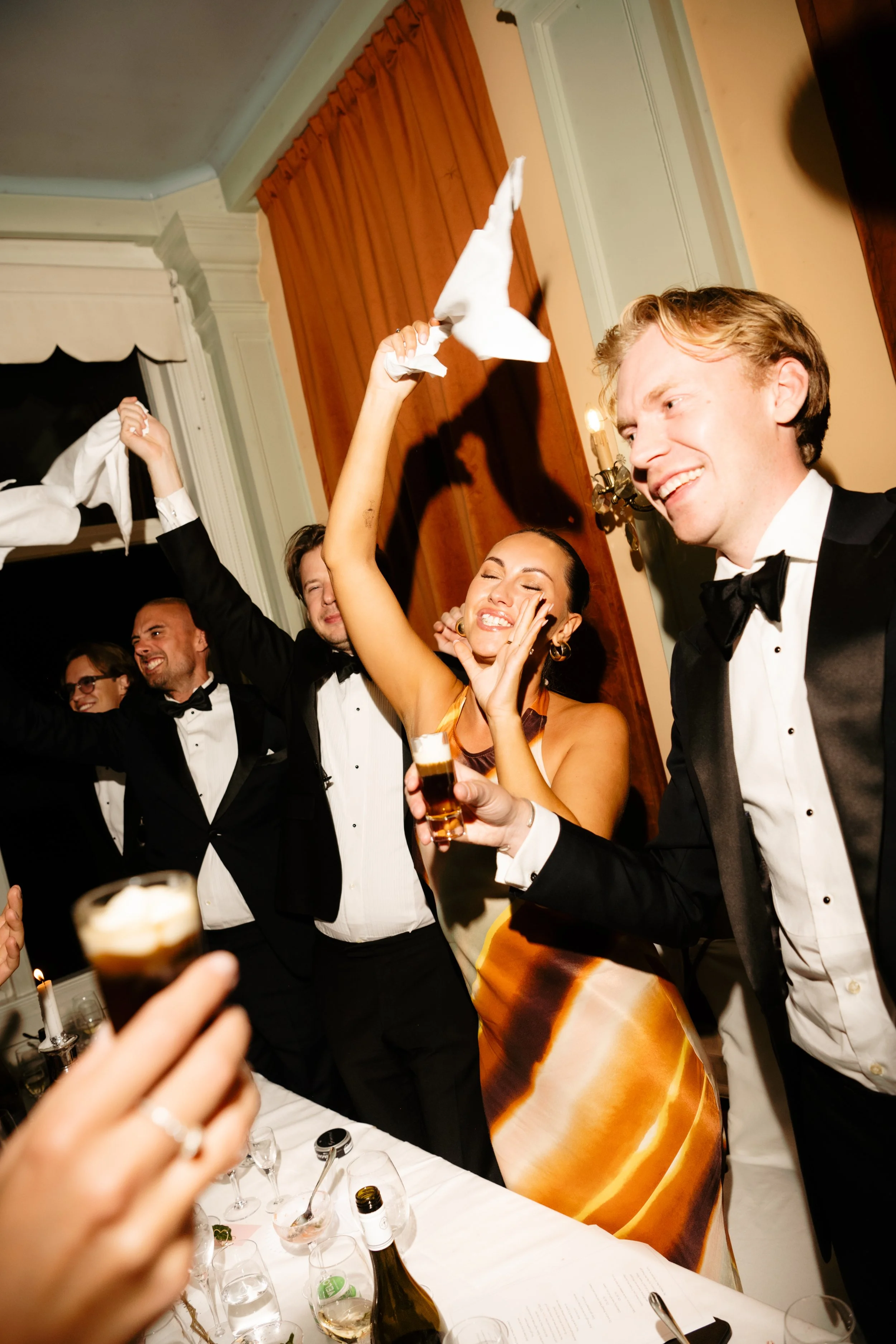 Celebration at a formal event with people in tuxedos and a woman in a colorful dress, smiling and dancing, holding napkins and glasses of drinks, with a table set with glassware and bottles in the foreground.