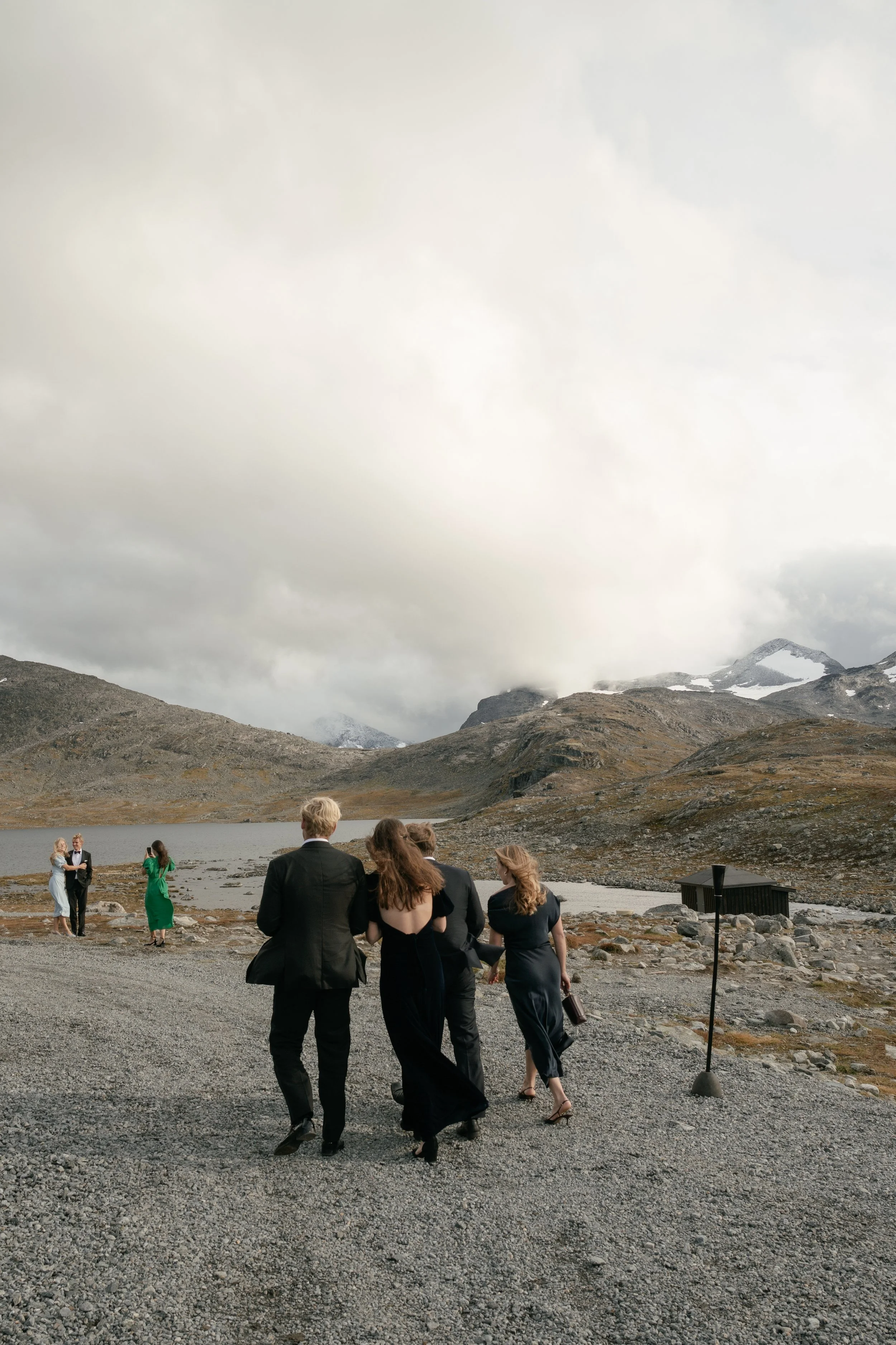 People dressed in formal attire walking along a gravel path near a lake with mountains in the background under a cloudy sky.