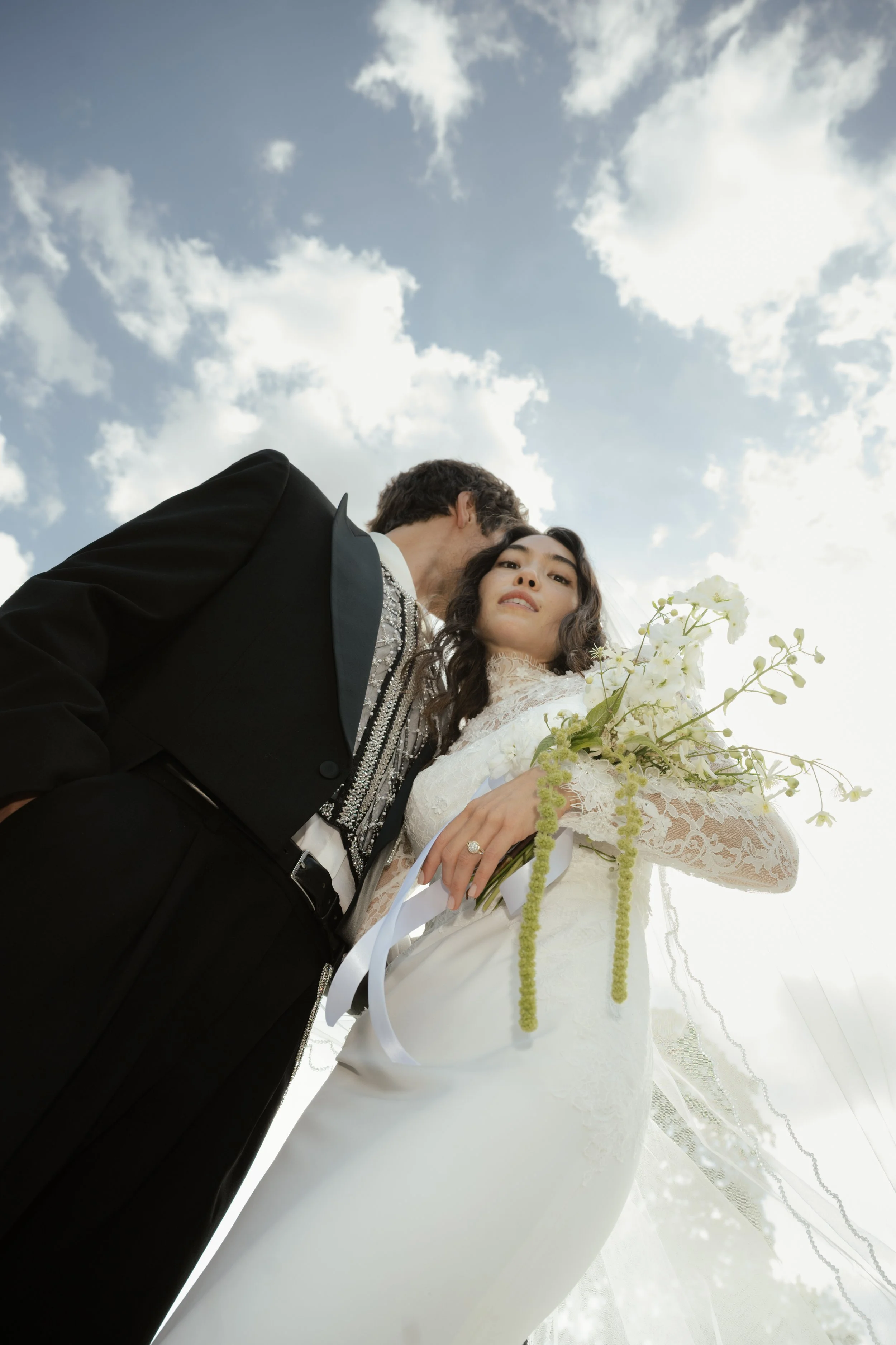 A bride and groom are standing close together outdoors against a bright sky with scattered clouds, dressed in formal wedding attire, with the bride holding a bouquet of white flowers.
