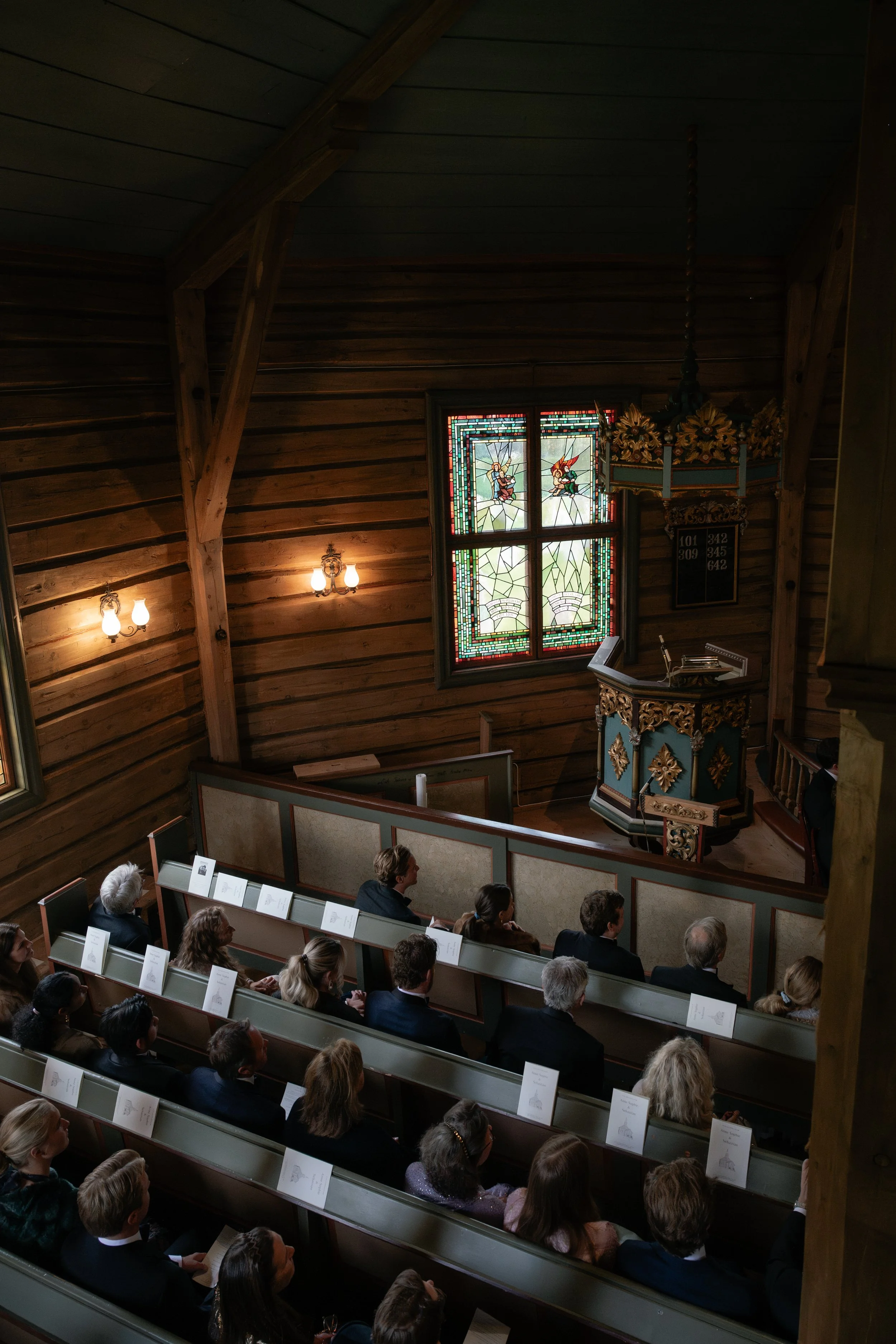 People attending a church service or event inside a wooden church, with stained glass windows and an ornate pulpit.