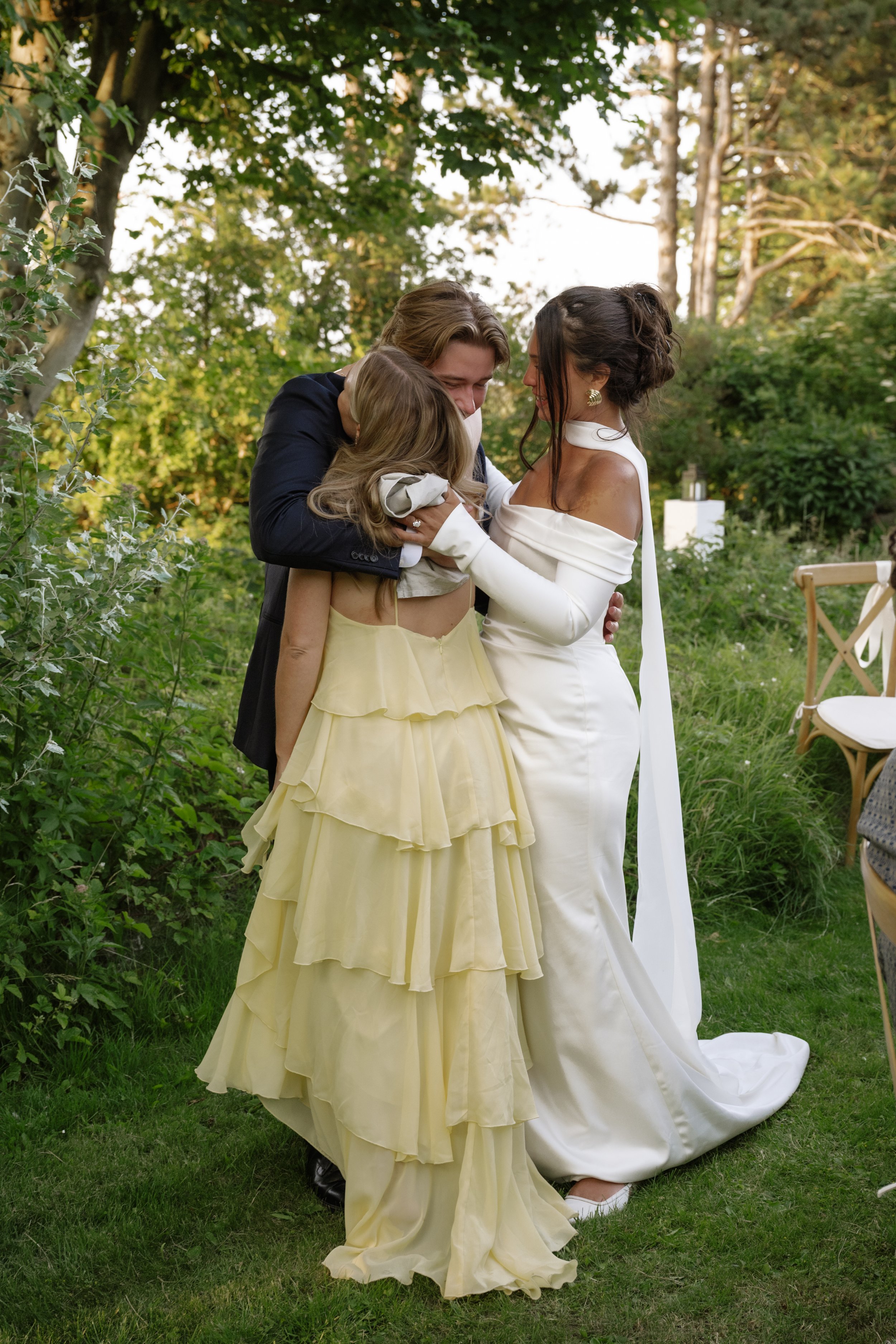 Three people, two women and a girl, sharing a happy embrace outdoors during a wedding or special event, with trees and greenery in the background.