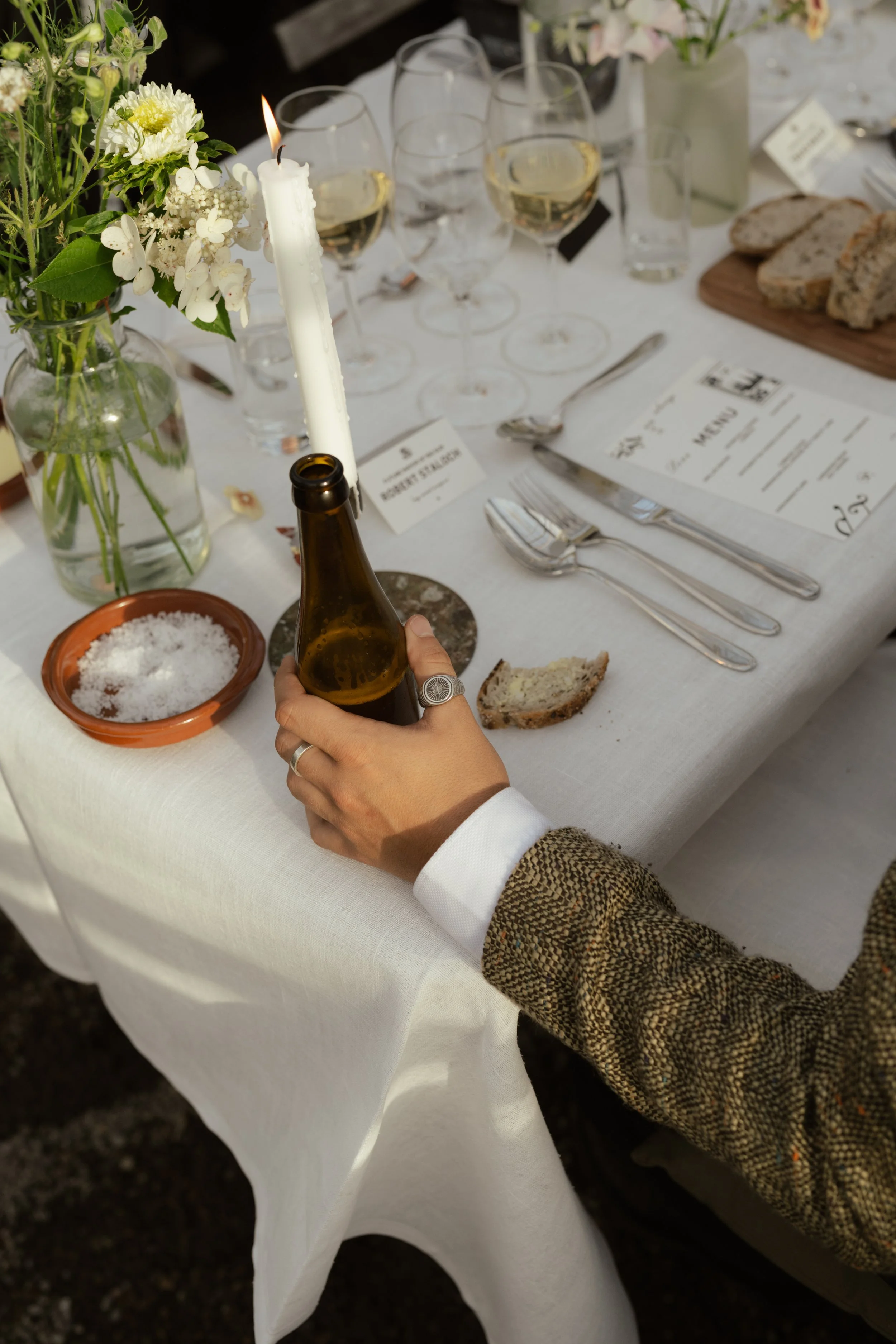 Person holding a beer bottle near a candle on a decorated dinner table with flowers, wine glasses, bread, and place cards.