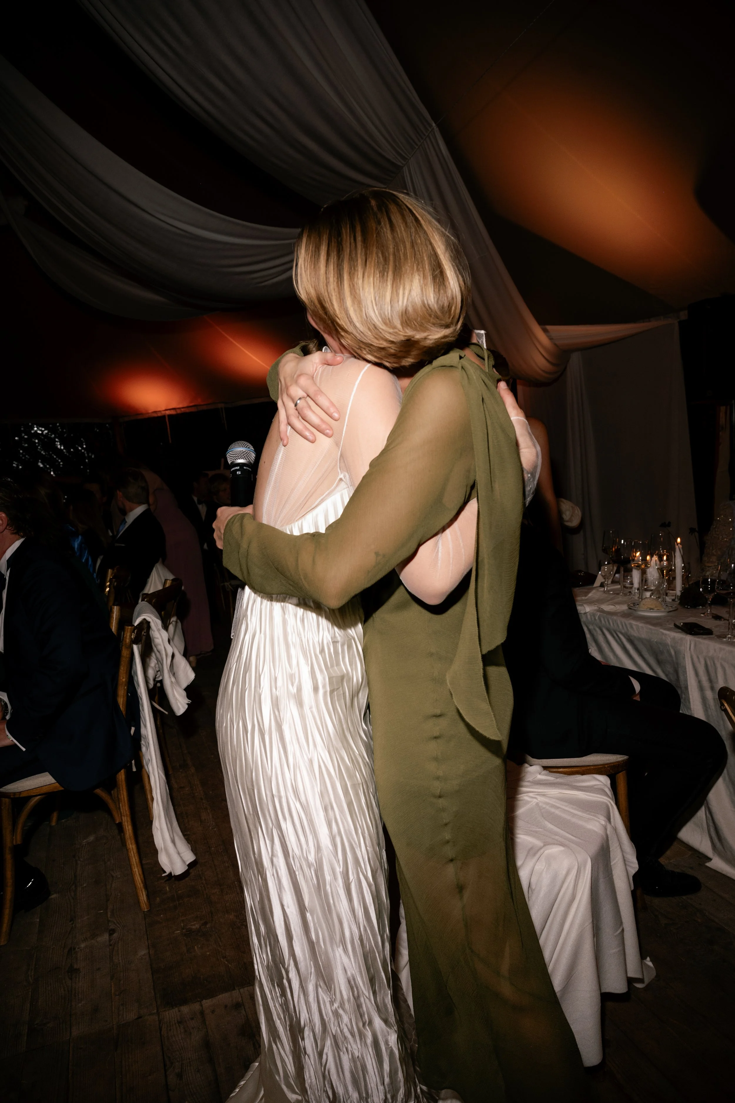 Two women hugging at a formal event, one wearing a white dress and the other in olive-green outfit, with a microphone in hand and a dinner table in the background.