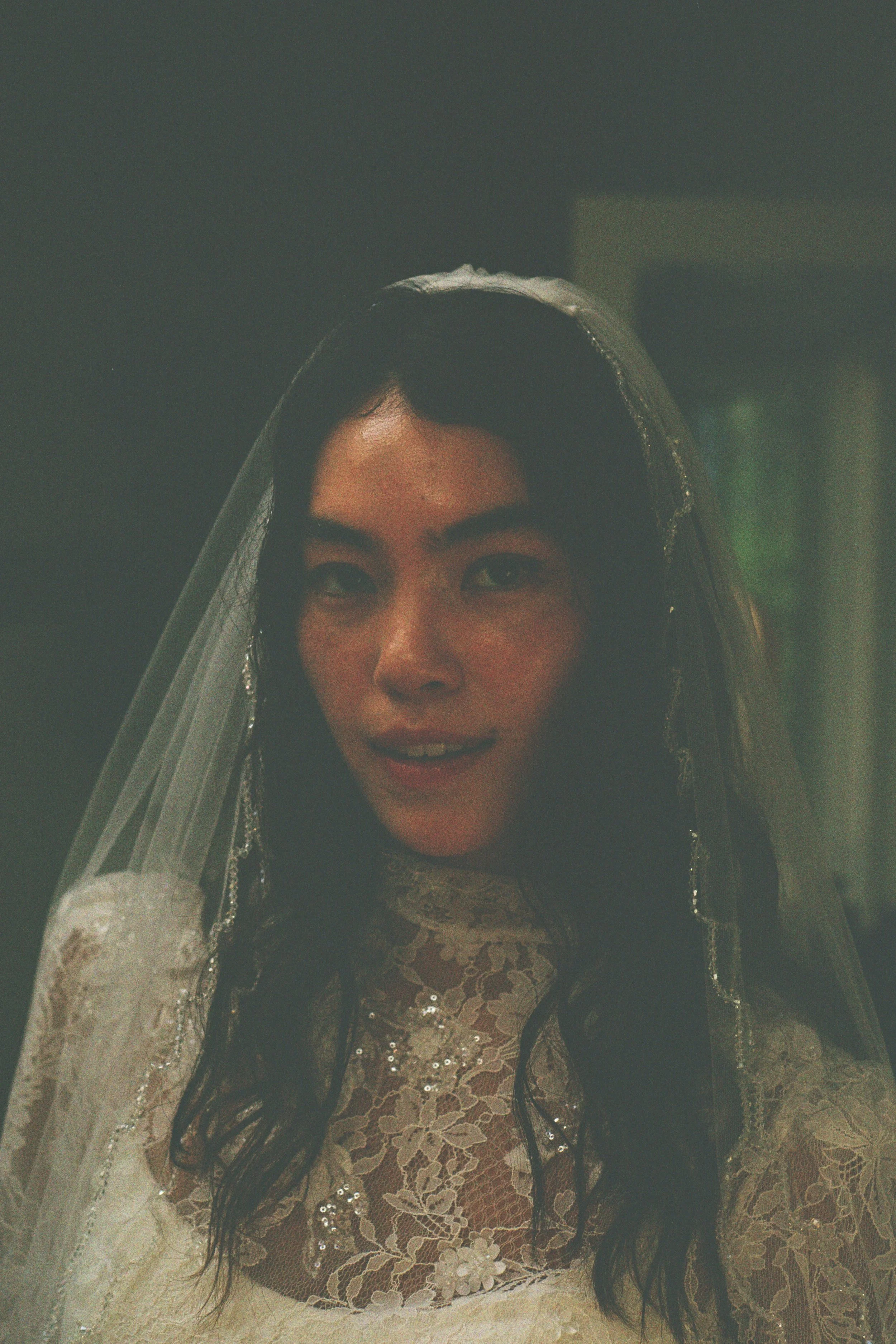 A woman dressed as a bride, wearing a wedding dress with lace and sequins, and a veil with intricate embellishments, smiling softly at the camera.