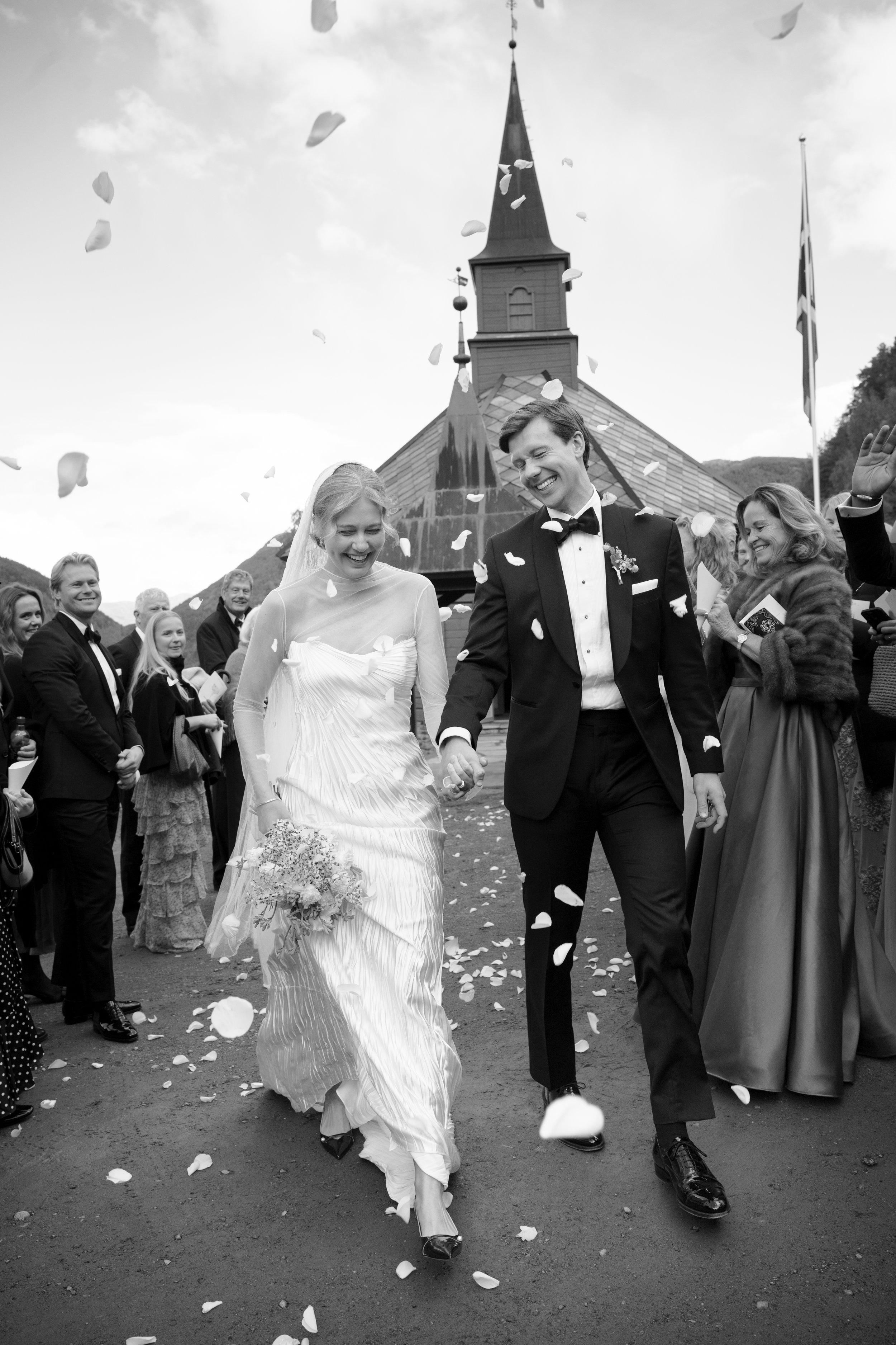 Black and white photo of a newly married couple smiling and holding hands, walking down the church steps while guests throw flower petals. The bride is in a white wedding dress holding a bouquet, and the groom is in a tuxedo. The scene is outdoors wi