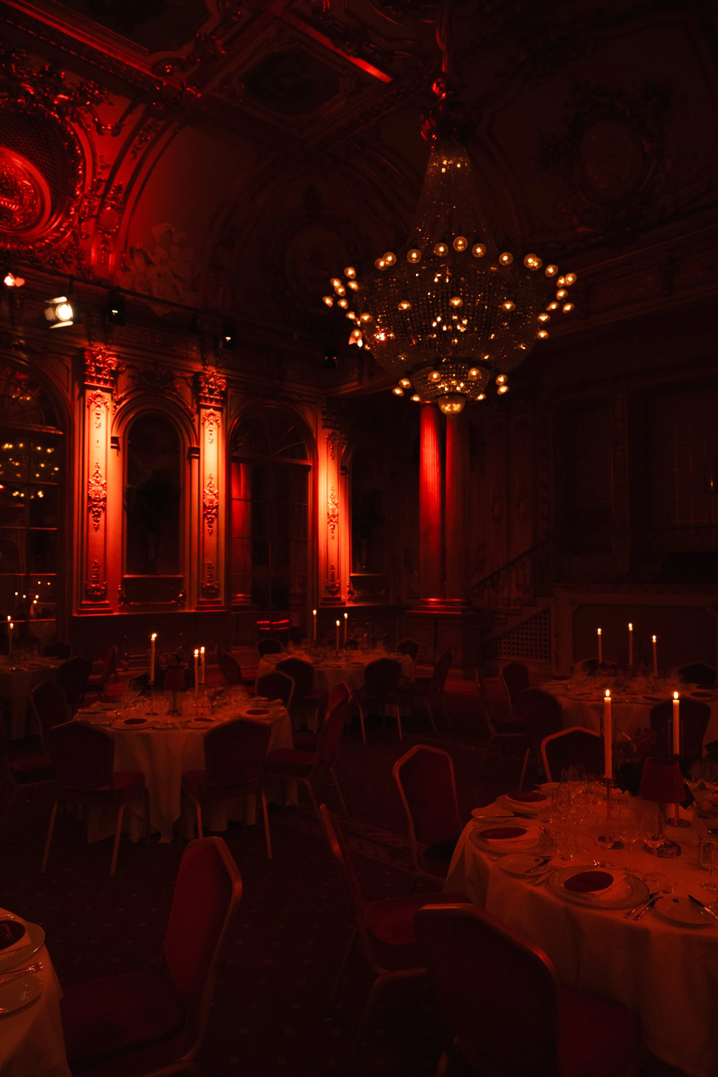 Elegant dining room with round tables set for a formal event, illuminated by candlelight and a large chandelier, with ornate walls and red lighting creating a luxurious atmosphere.