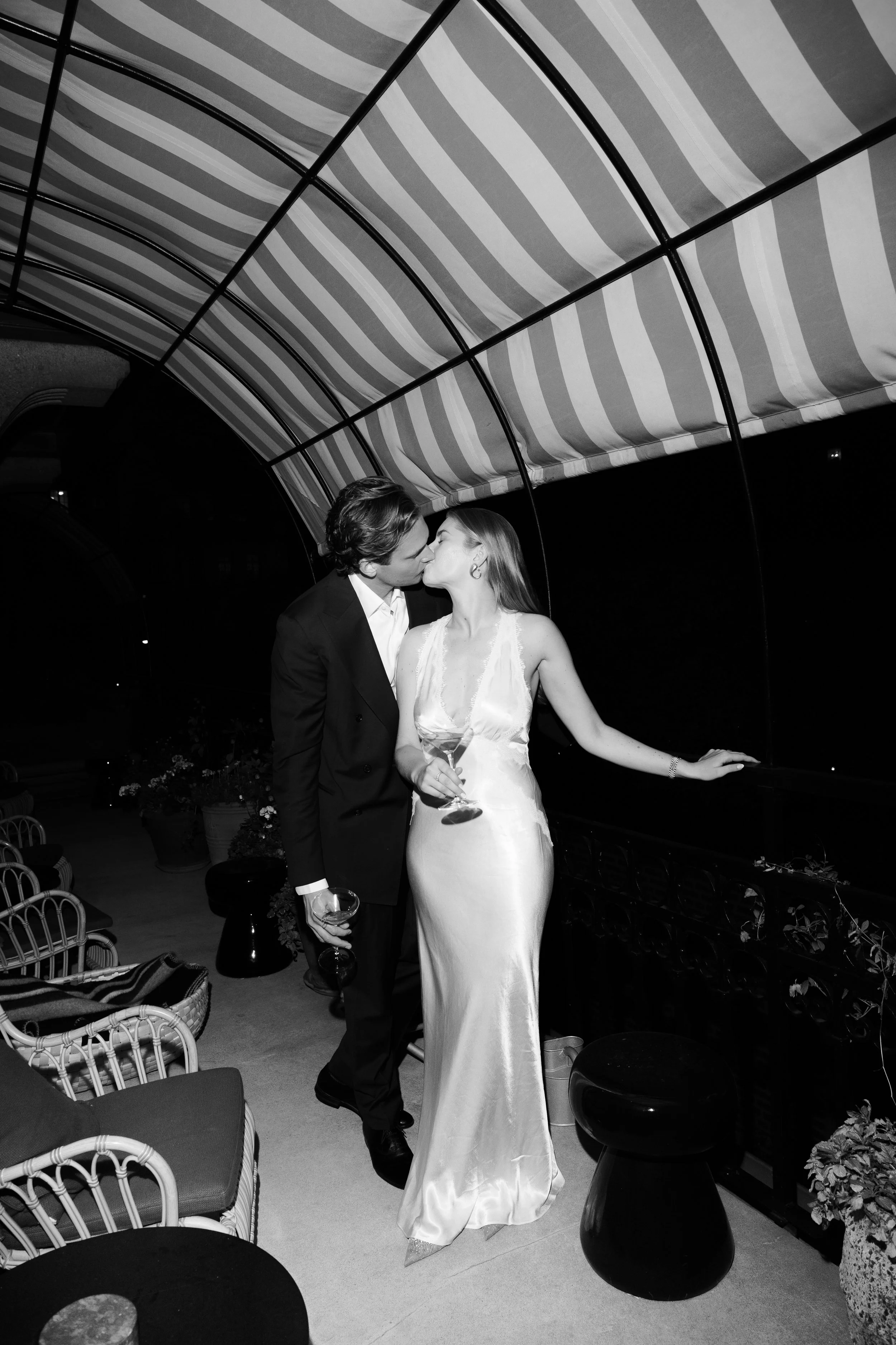 A black and white photo of a couple kissing on a balcony at night. The woman wears a satin dress, holds a glass of wine, and leans on the railing. The man wears a suit and also holds a wine glass. There are chairs, a small stool, and potted plants on