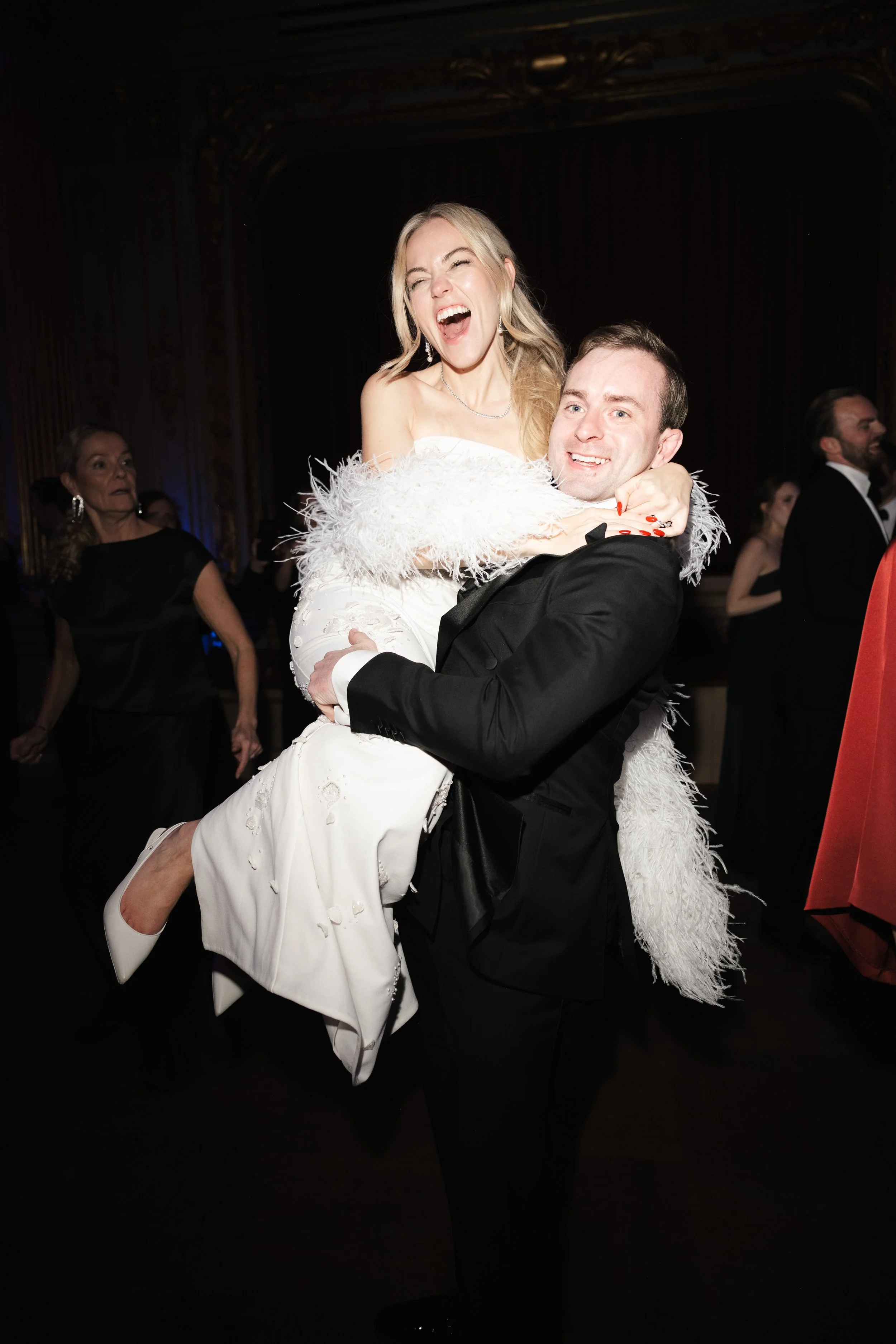 A man in a black tuxedo lifts a woman in a white dress with feathered sleeves at a formal event, both smiling and appearing joyful, with other guests dancing in the background.