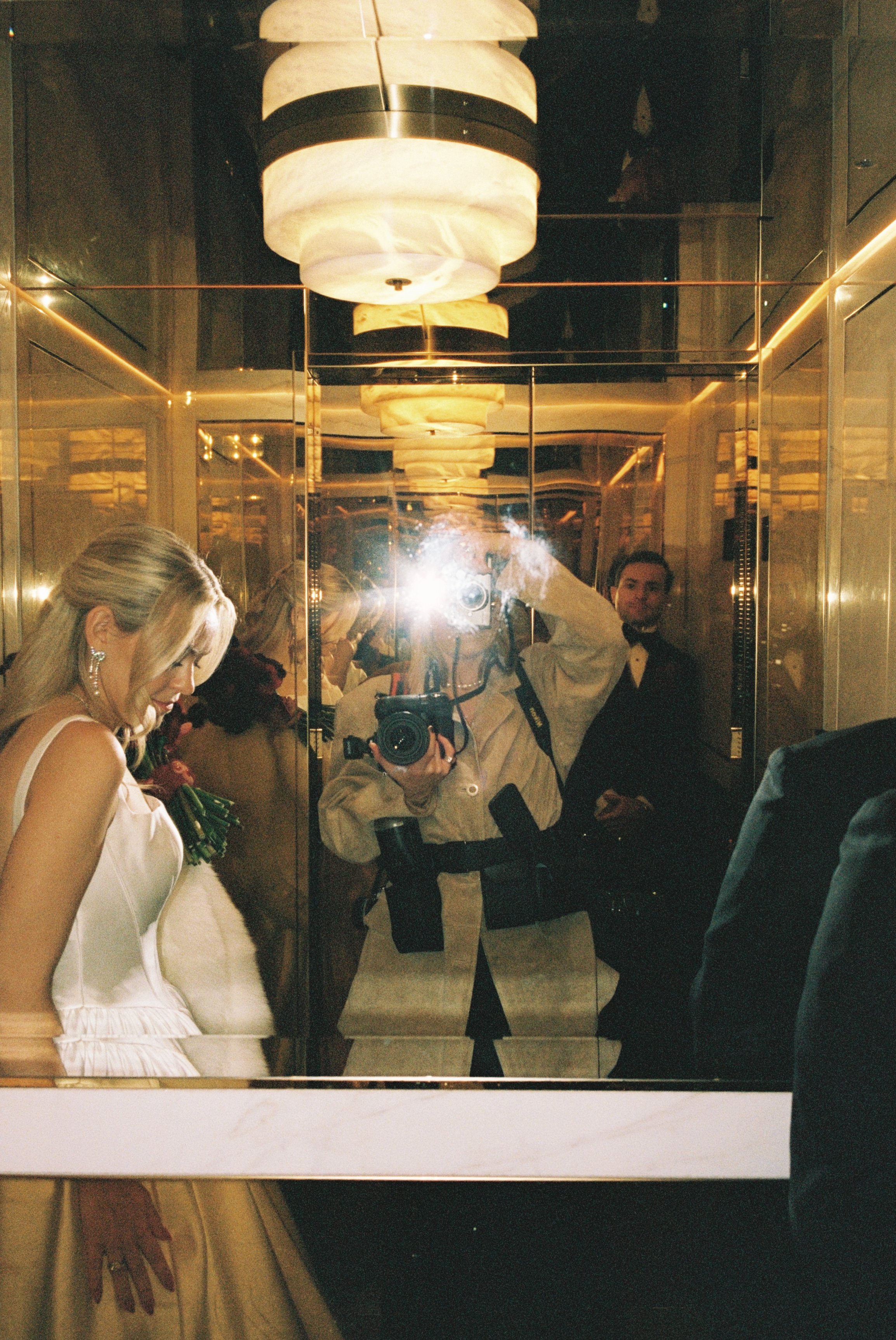 A woman in a white dress is in an elevator surrounded by mirrors. A man in a black tuxedo stands behind her. The photographer’s flash is reflected in the mirror, capturing the scene.