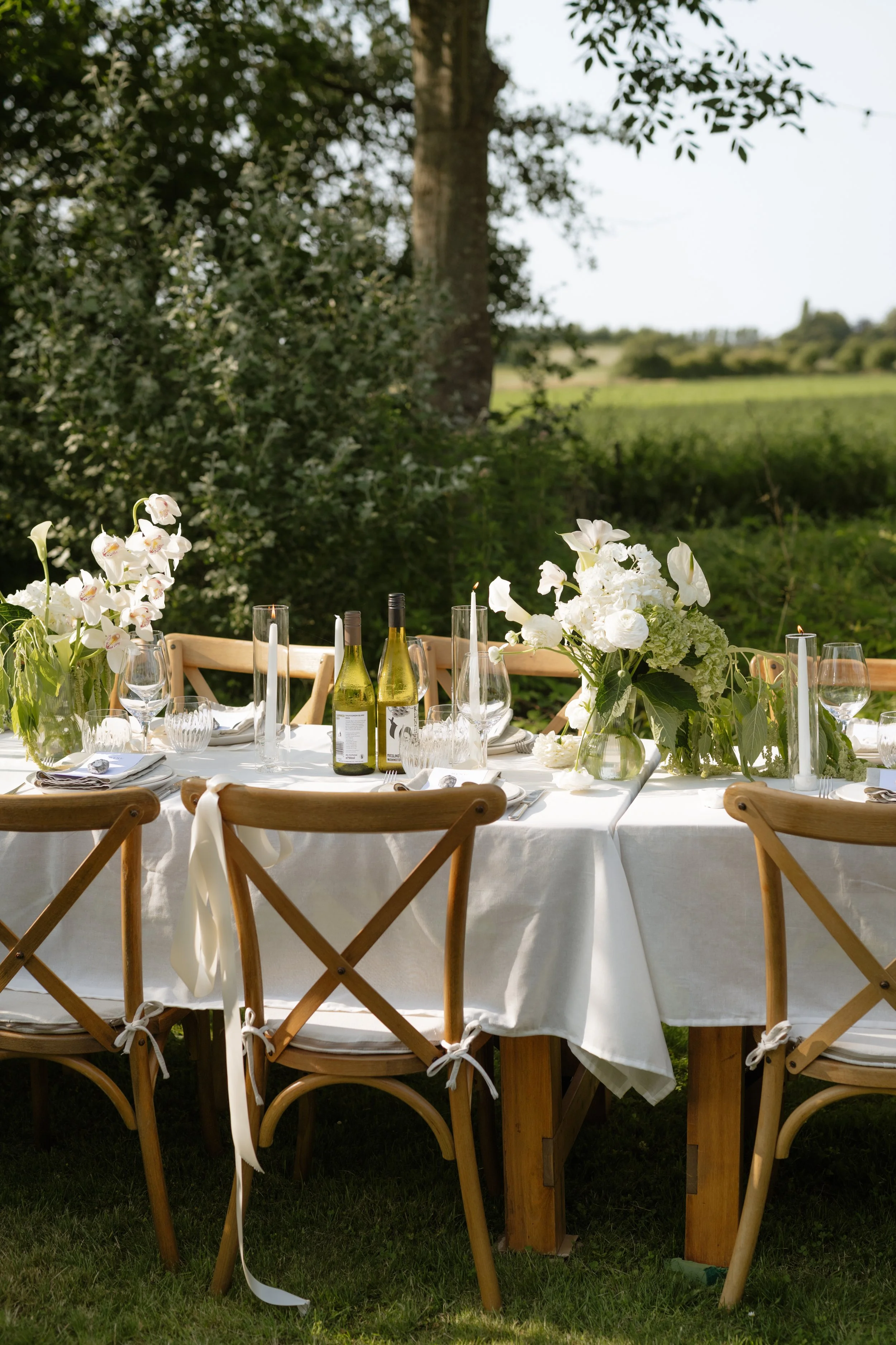 A beautifully set outdoor dining table with white tablecloth, floral centerpieces, wine bottles, and glassware, situated in a lush green garden with trees and fields in the background.