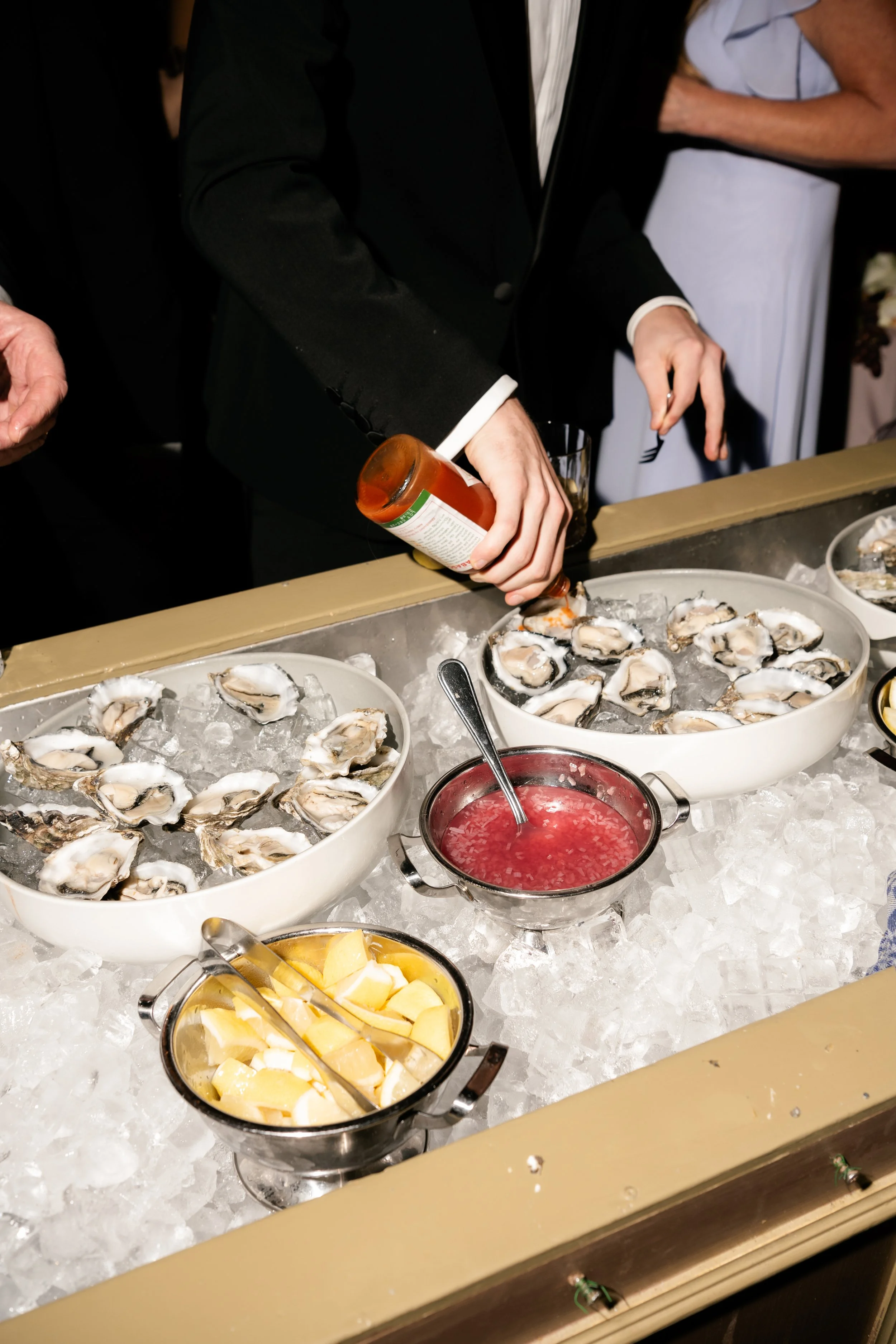 Person in a black suit pouring ketchup over a bowl of oysters on ice, with bowls of lemon wedges and cocktail sauce nearby.