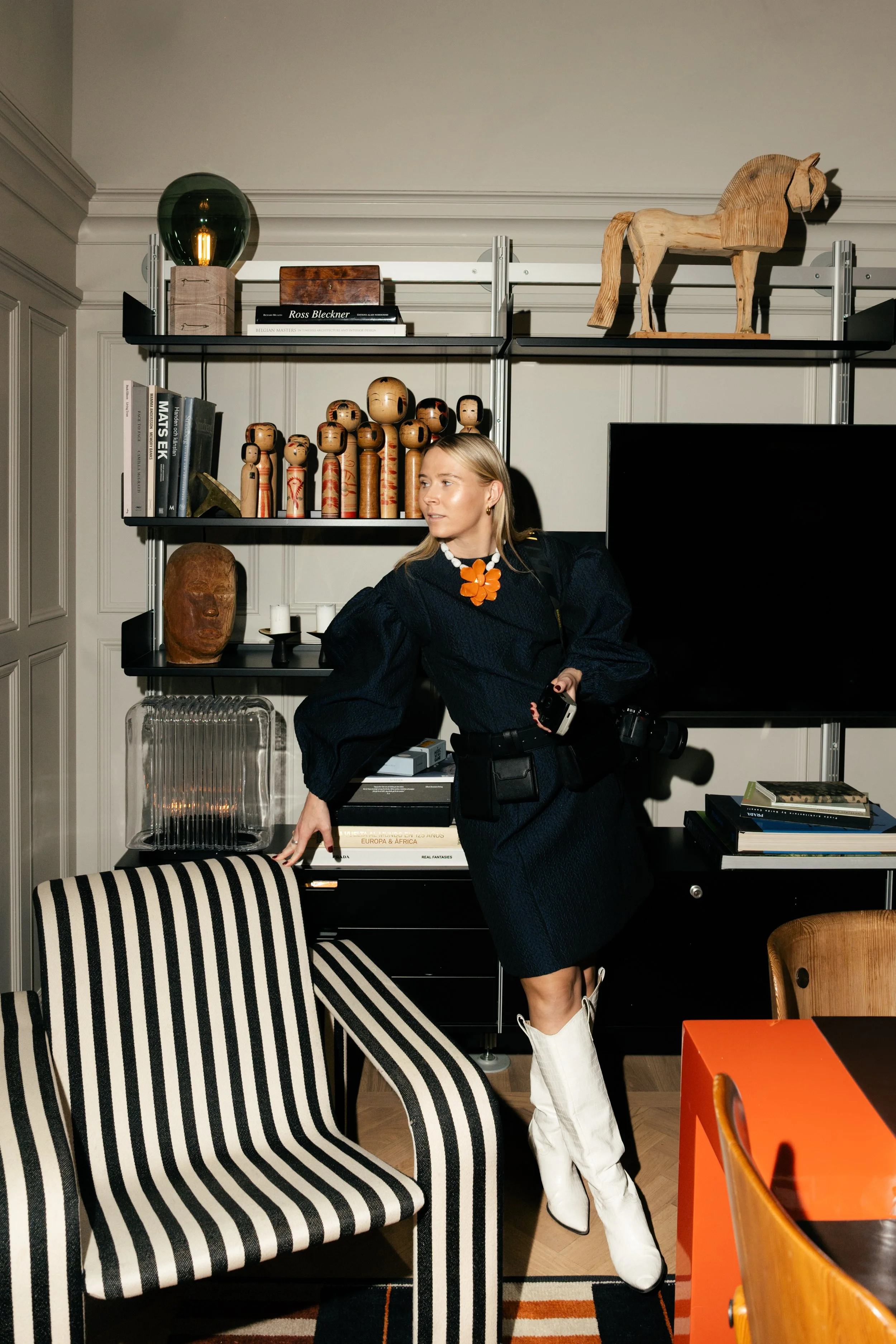 A woman with blonde hair wearing a black dress with puffy sleeves, white high boots, and a large orange flower necklace, standing in a modern living room next to a striped armchair and black shelving filled with decorative objects and books.