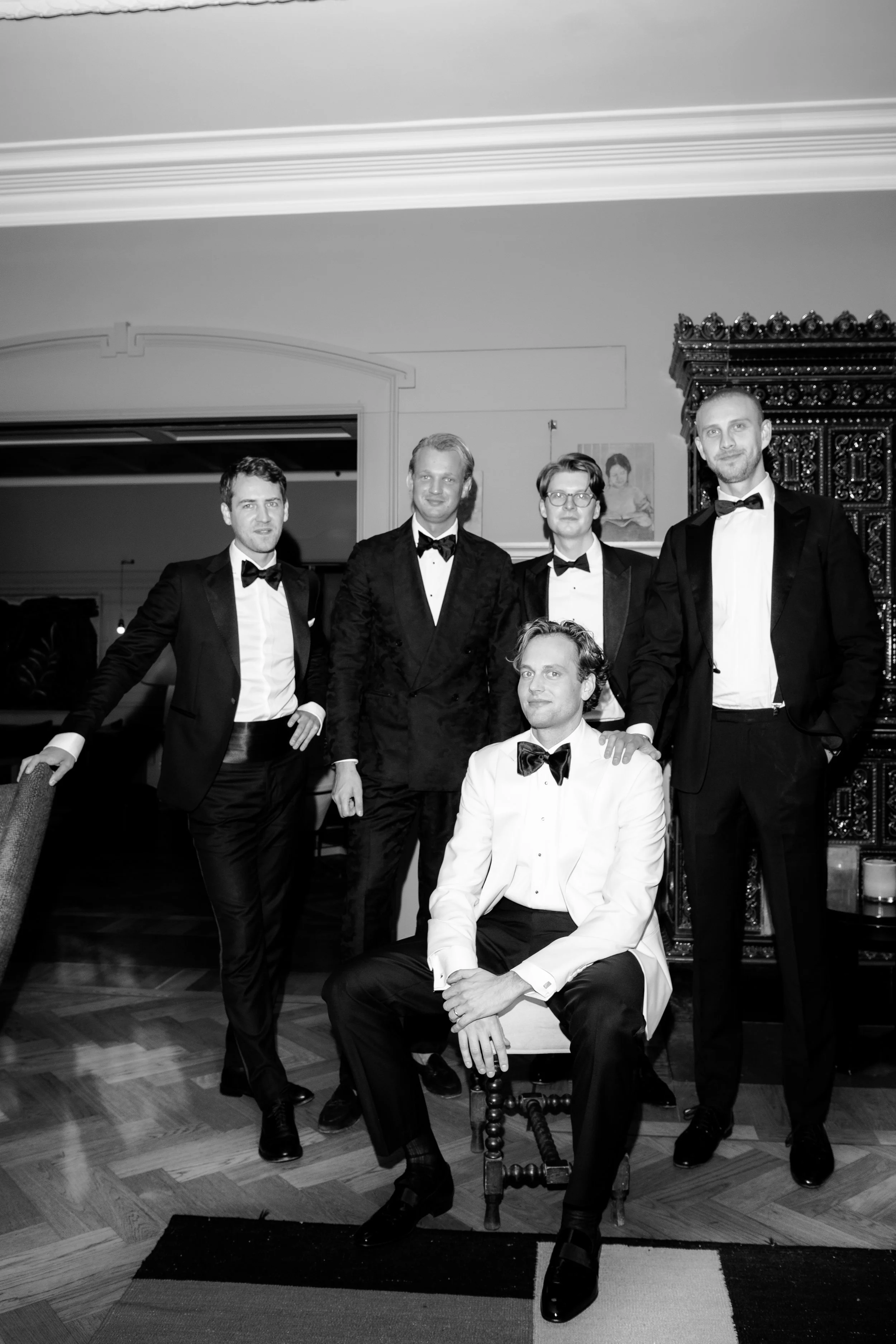 A group of six men dressed in tuxedos posed together indoors, with one seated in front and the others standing behind him, at a formal event or celebration.