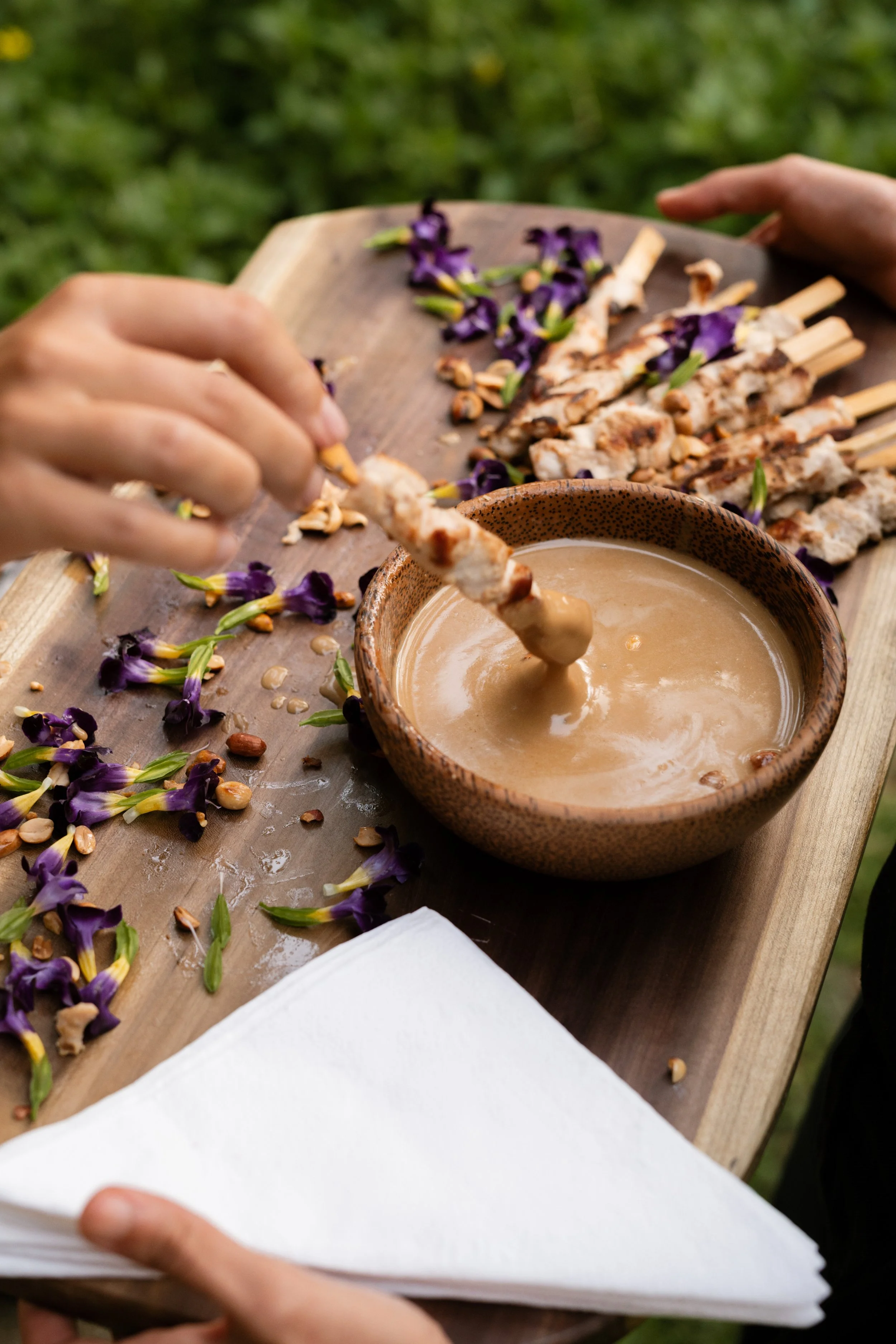 Person serving skewered grilled chicken into a bowl of peanut sauce on a wooden serving platter decorated with purple flowers and peanuts, with a white napkin at the corner.