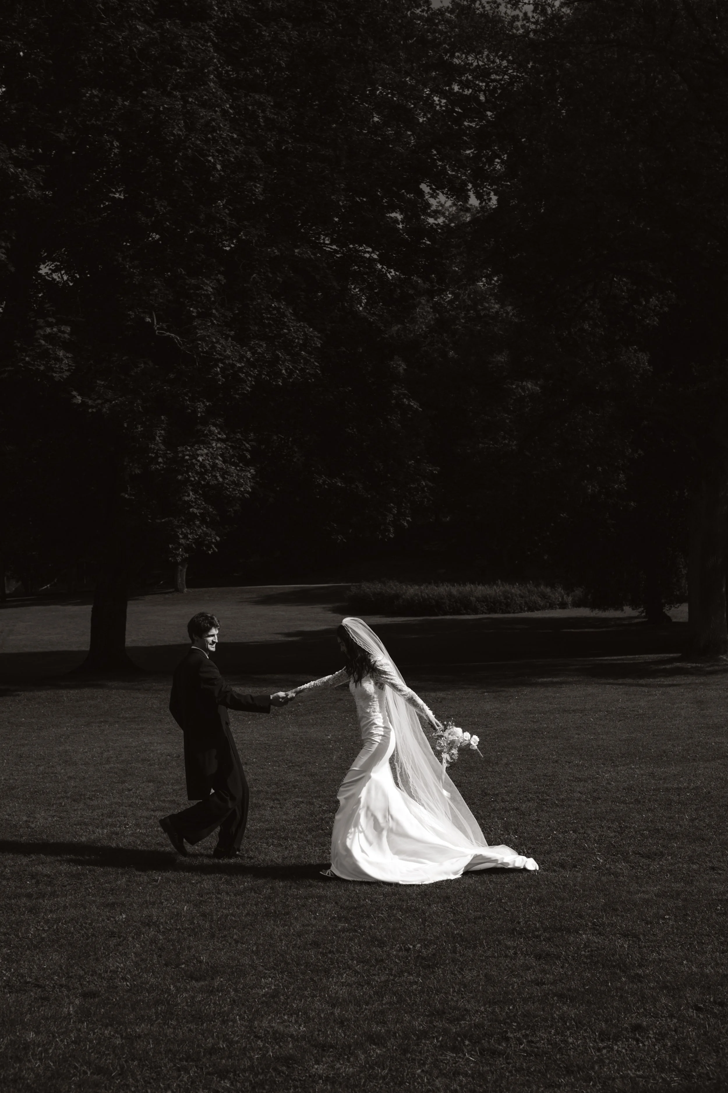 A black and white photo of a bride and groom holding hands and dancing outdoors on a grassy field, with large trees in the background.