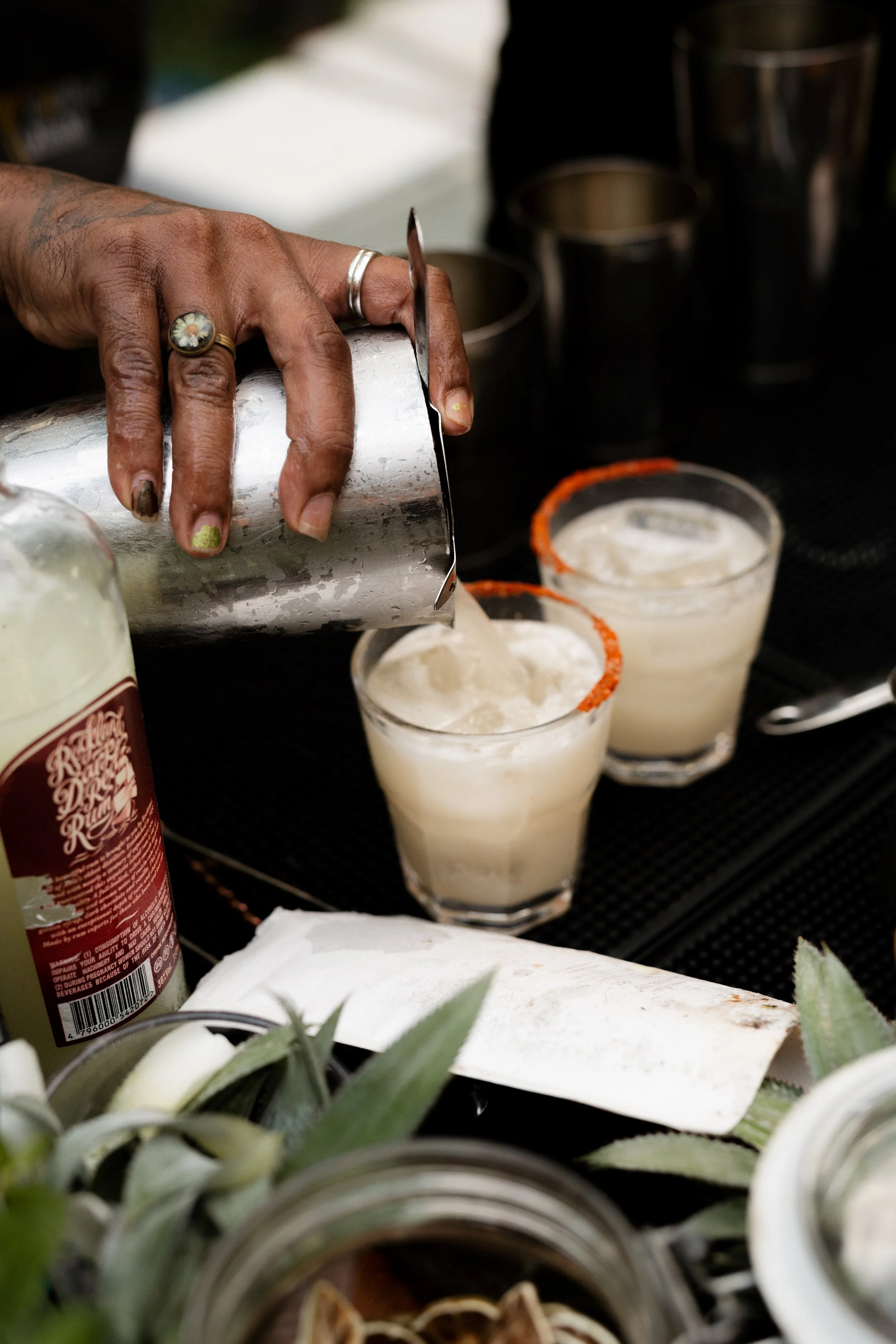 Bartender pouring a cocktail into glasses with salted rims, surrounded by bar supplies and plants.