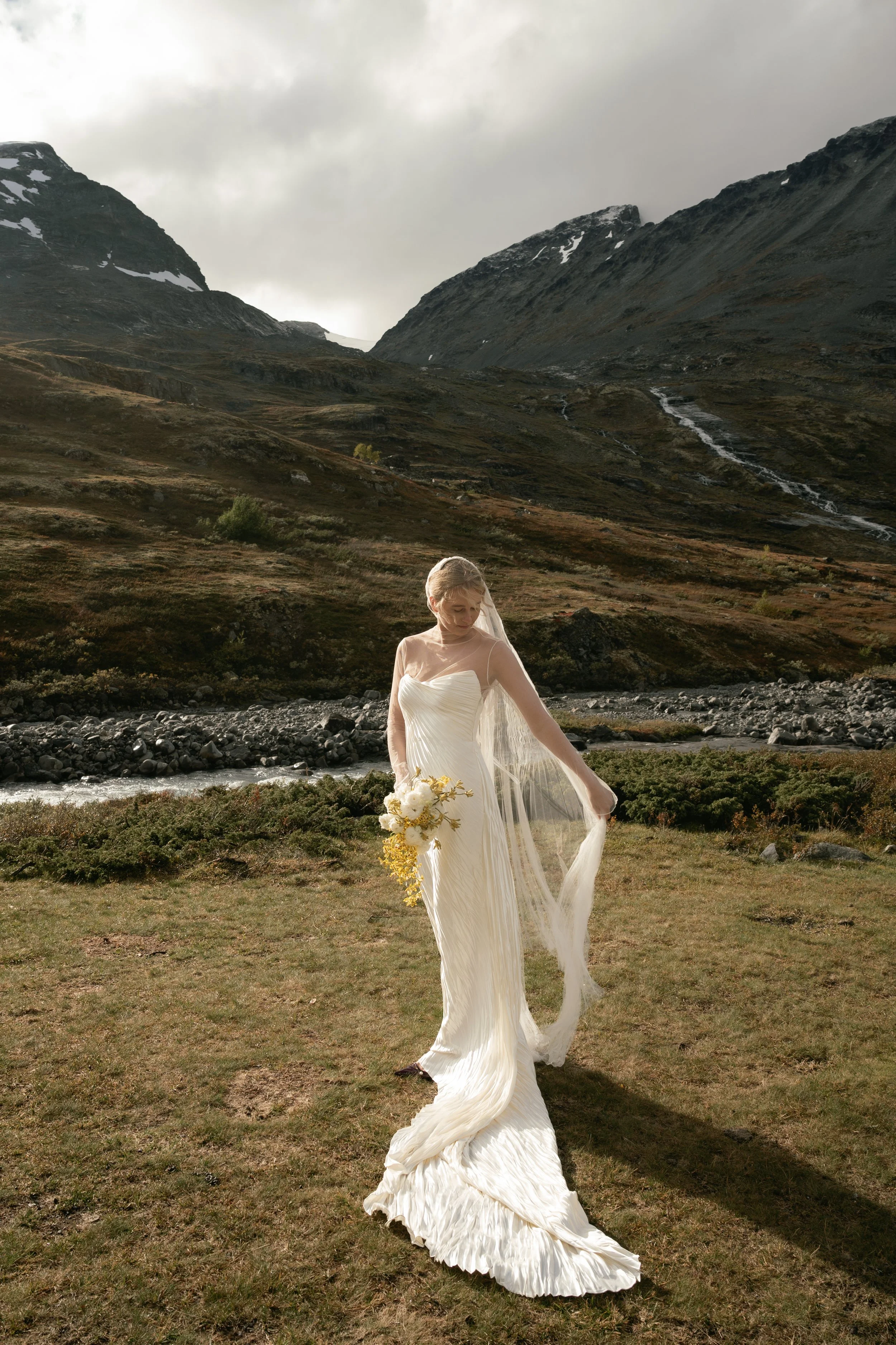 A bride in a white wedding dress standing on grass in a mountainous landscape with a river behind her. She holds a bouquet of white and yellow flowers and looks down, with her veil flowing to one side.
