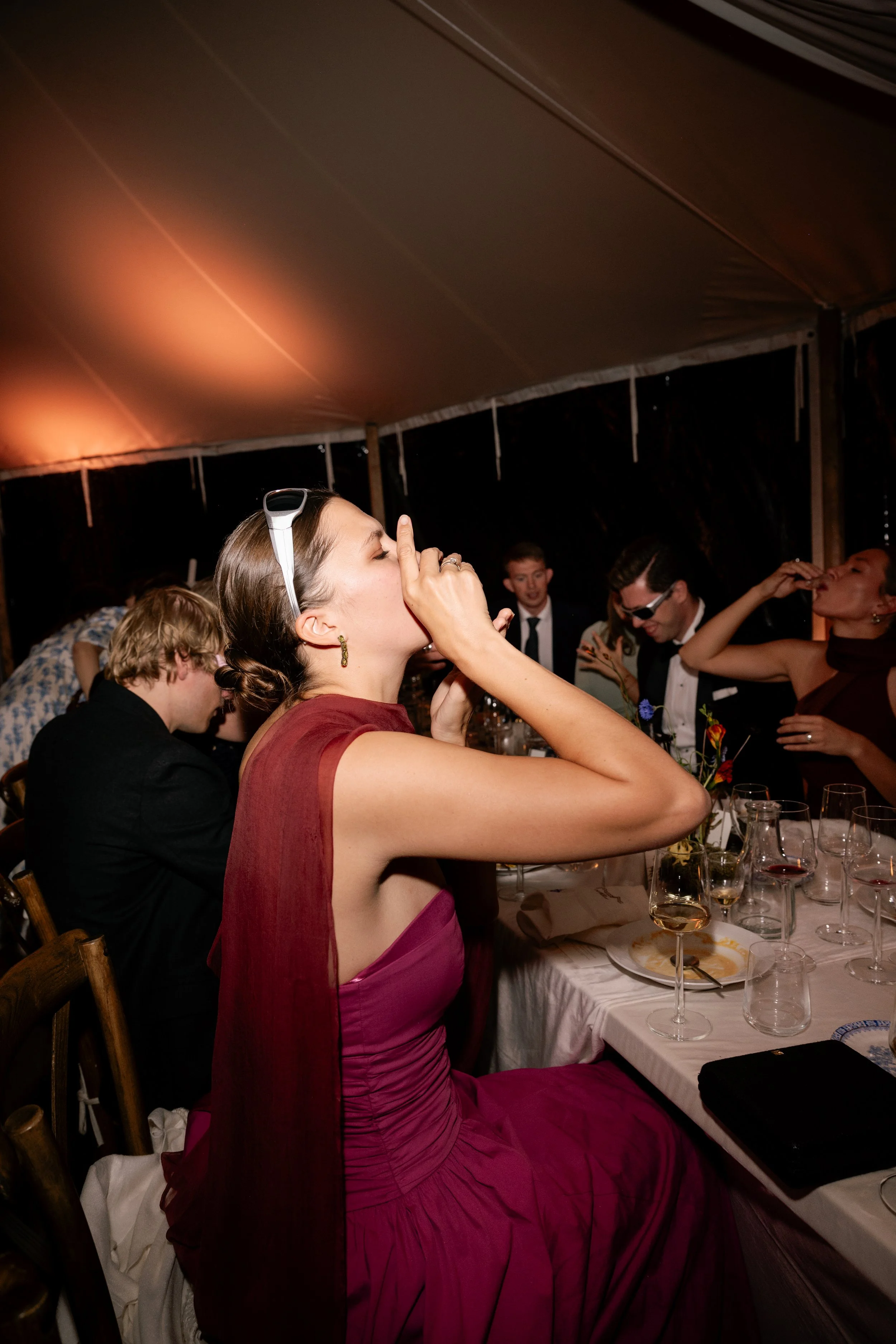 A woman in a burgundy dress at a dinner party is taking a shot of alcohol, with other guests in the background inside a tent with warm lighting.