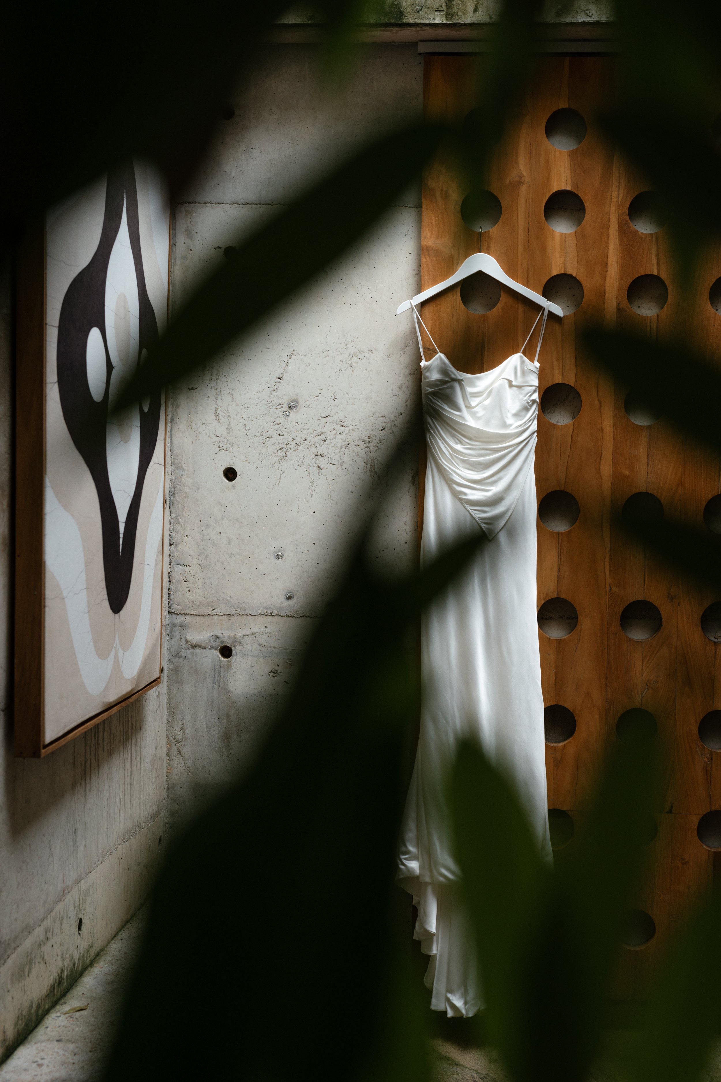 White wedding dress hanging on a white hanger against a textured concrete wall with a decorative wooden panel and an abstract artwork partially obscured by leaves in the foreground.
