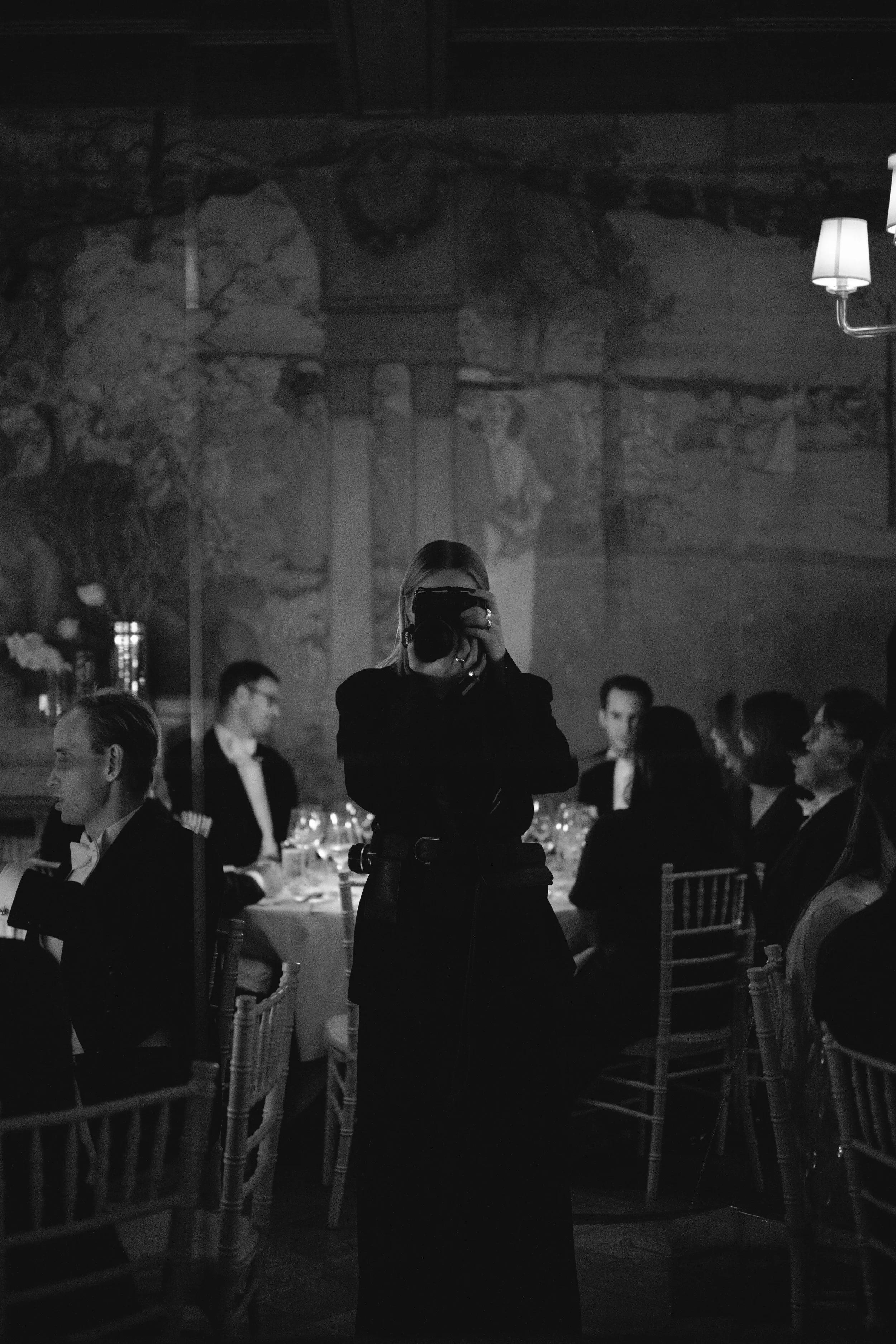 A woman taking a photo in a dimly lit restaurant with people seated at tables, some in tuxedos, and a mural on the wall behind.
