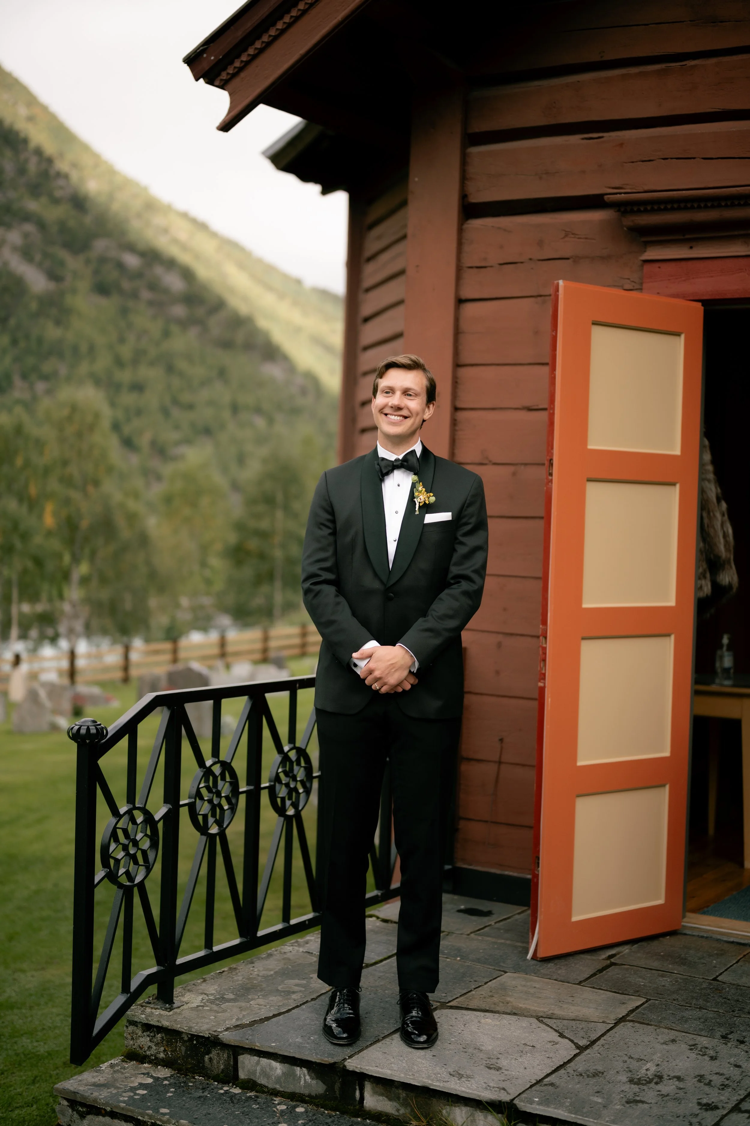 A man in a tuxedo standing outside a rustic wooden building, smiling with mountains and trees in the background.