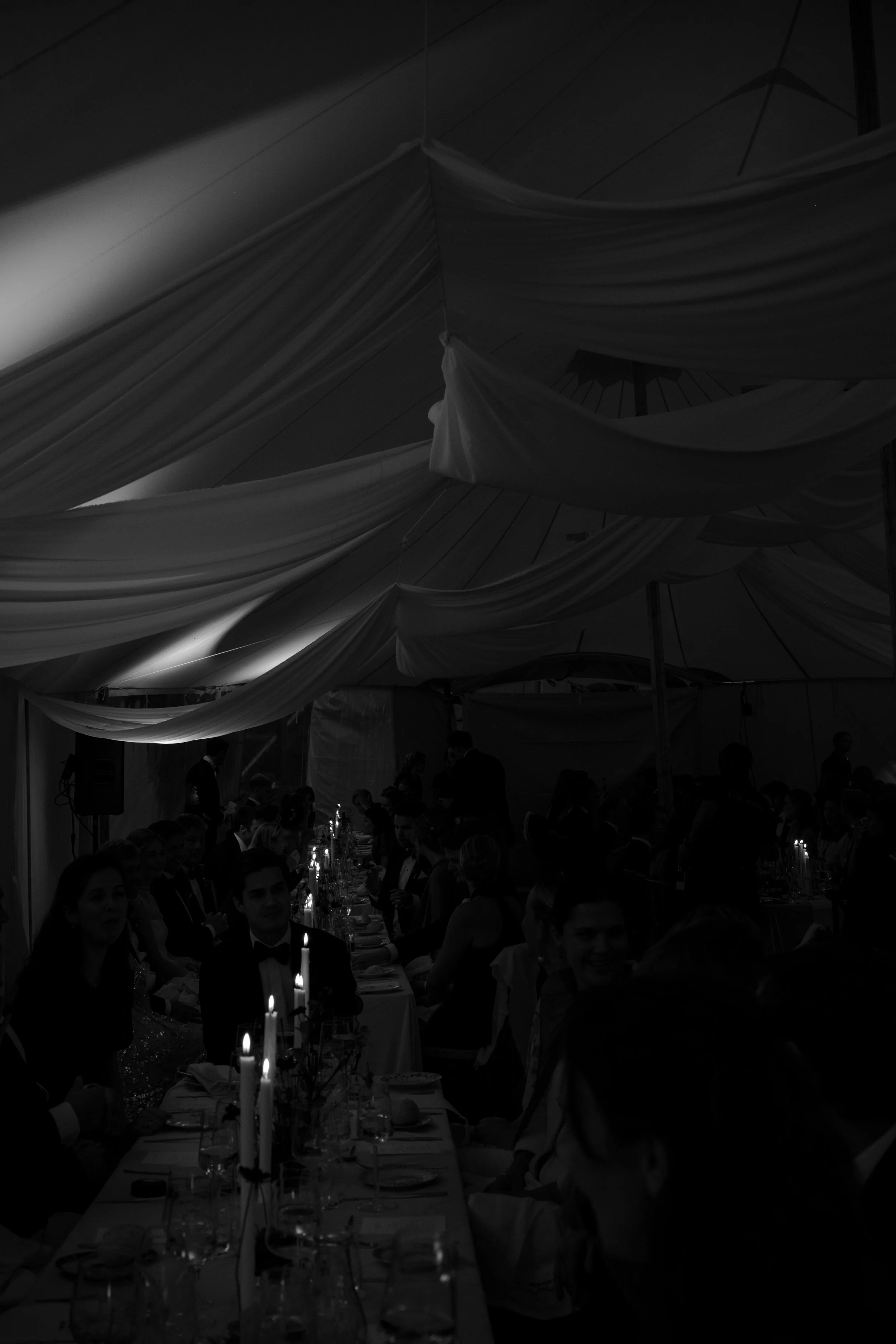 A black and white photo of a formal dinner event with guests seated at a long table, decorated with lit candles and elegant tableware inside a tent with draped fabric ceiling.