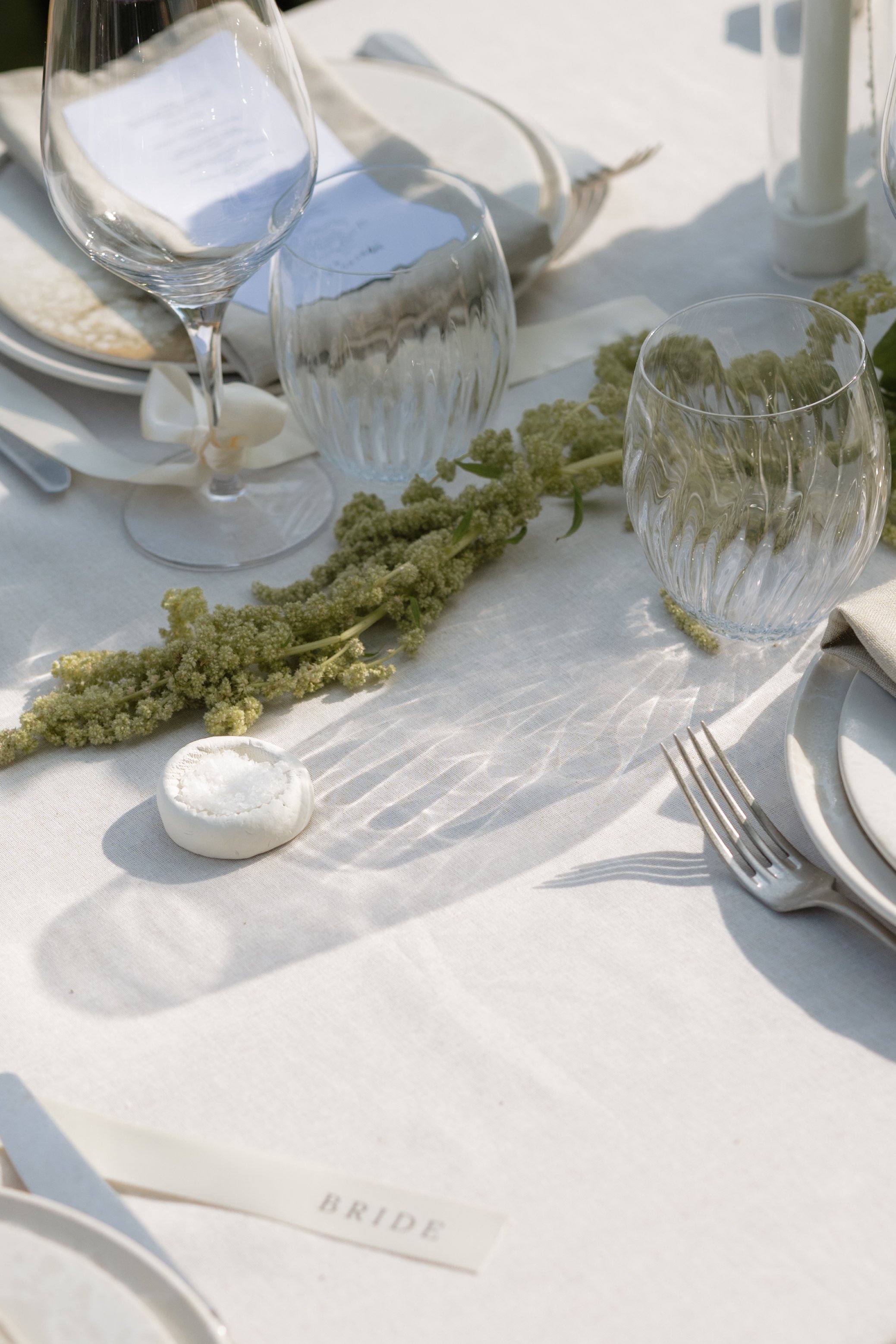 Elegant wedding table setting with clear wine and water glasses, white plates, a white napkin, a small white candle, and a green floral centerpiece on a white tablecloth.