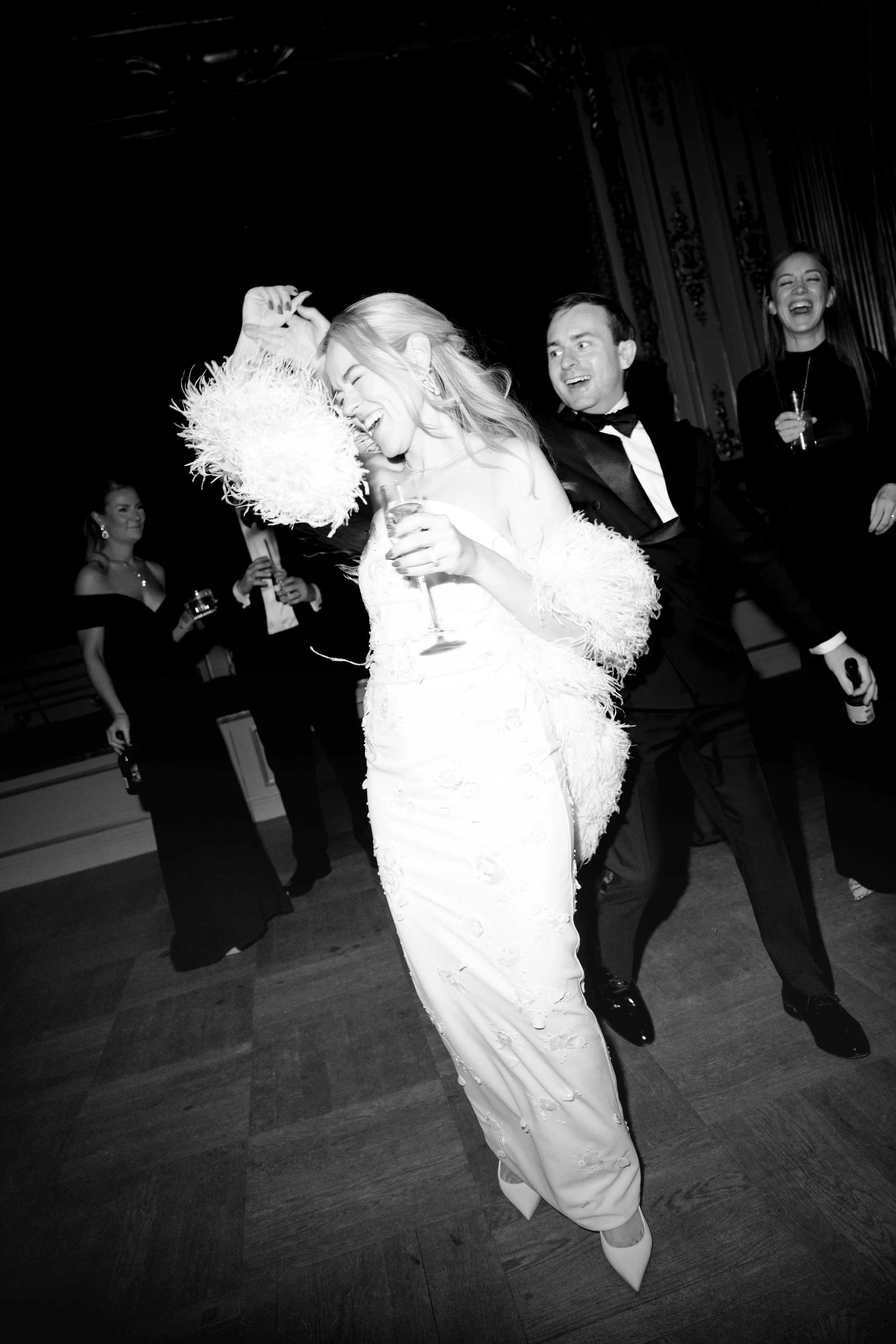 People dancing and celebrating at a formal event, with women in elegant dresses and men in tuxedos, holding drinks and smiling.