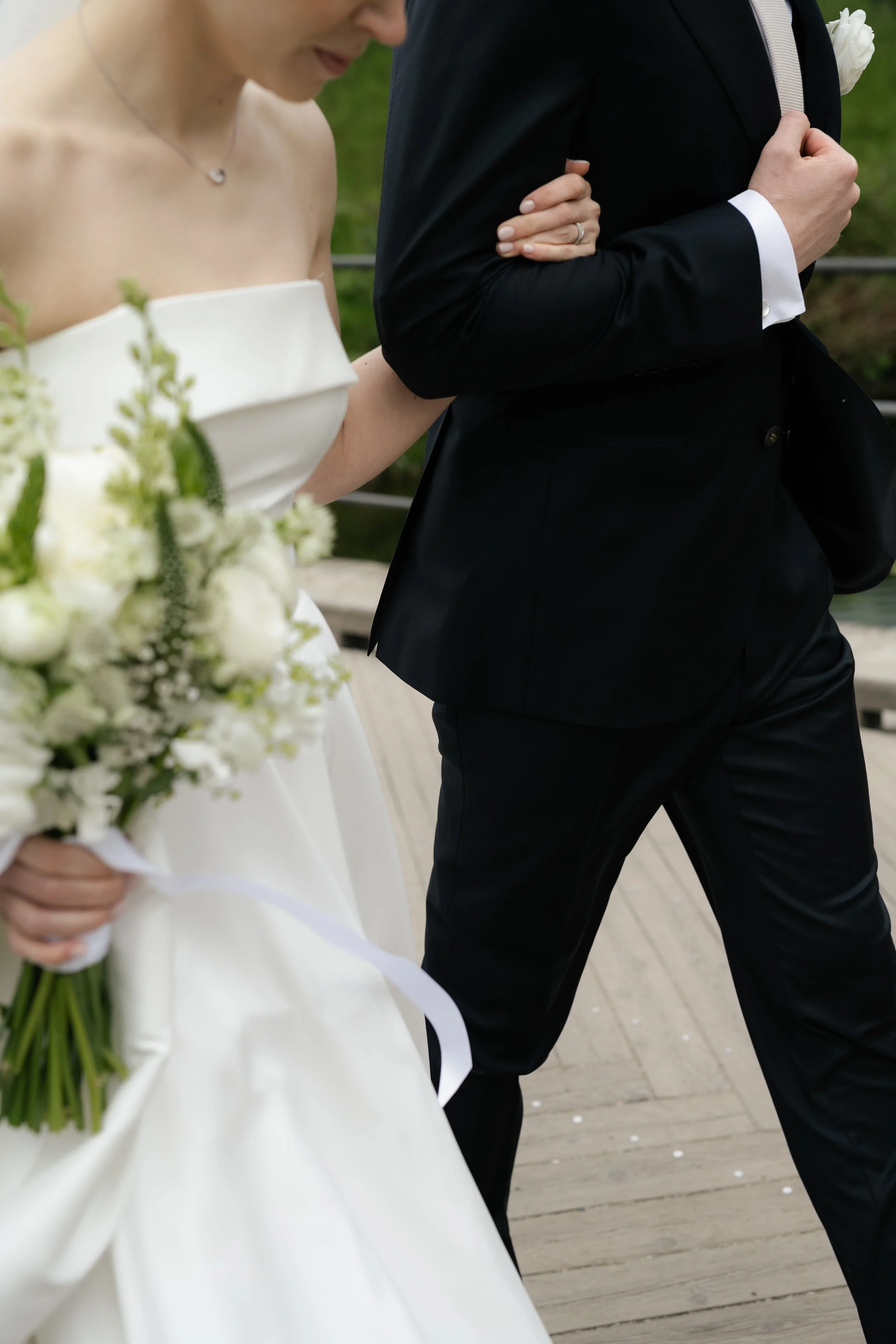 A couple dressed in wedding attire, with the bride holding a bouquet of white flowers and the groom wearing a black tuxedo, walking together on a wooden pathway.