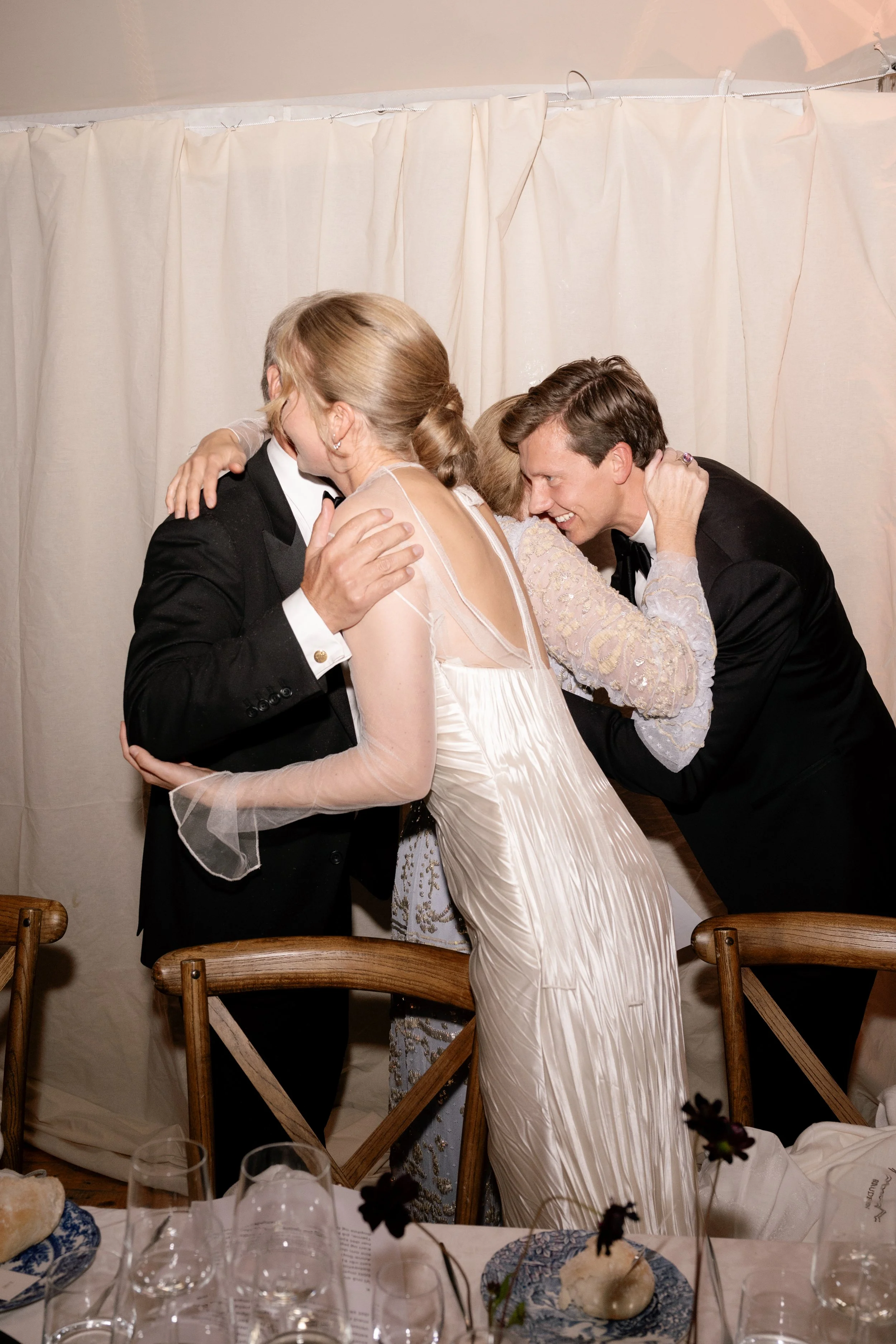 Group of people hugging at a celebration or wedding with a cream-colored curtain backdrop and a table with wine glasses and plates in the foreground.