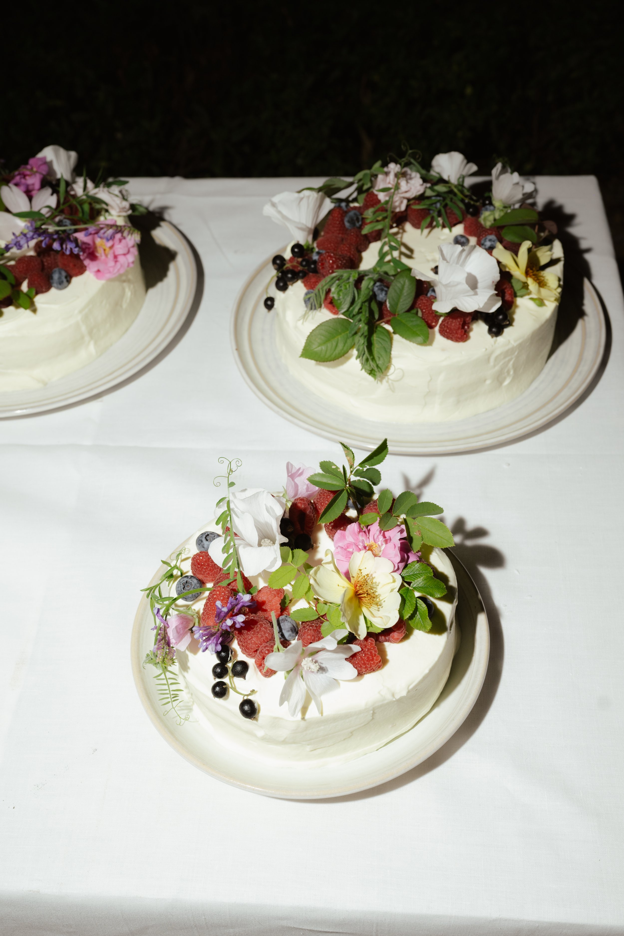 Three cakes decorated with fresh berries, flowers, and green leaves on a white tablecloth
