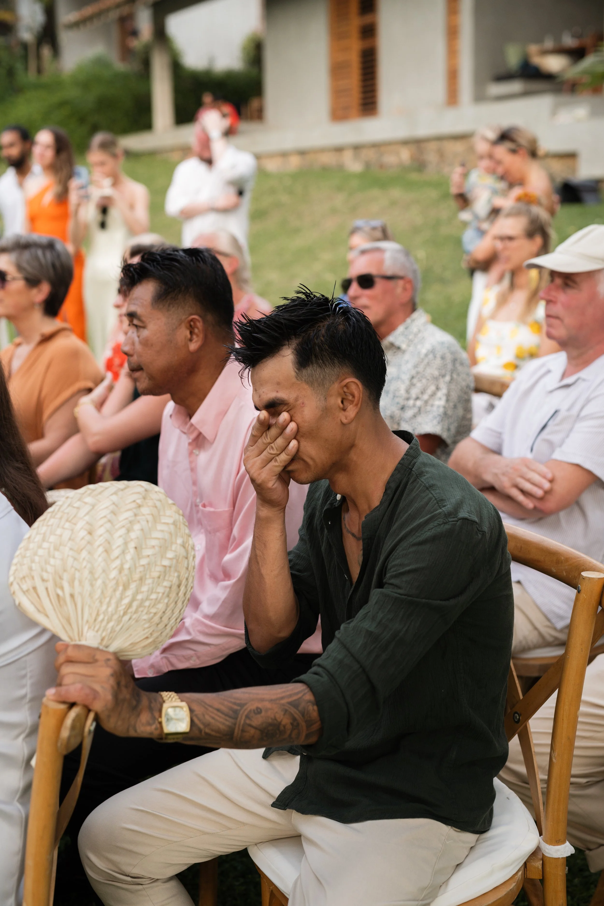 A group of people attending an outdoor event, with some individuals visibly emotional, including a man in dark clothing holding a fan and wiping his eye, and others sitting and standing in the background near a building.