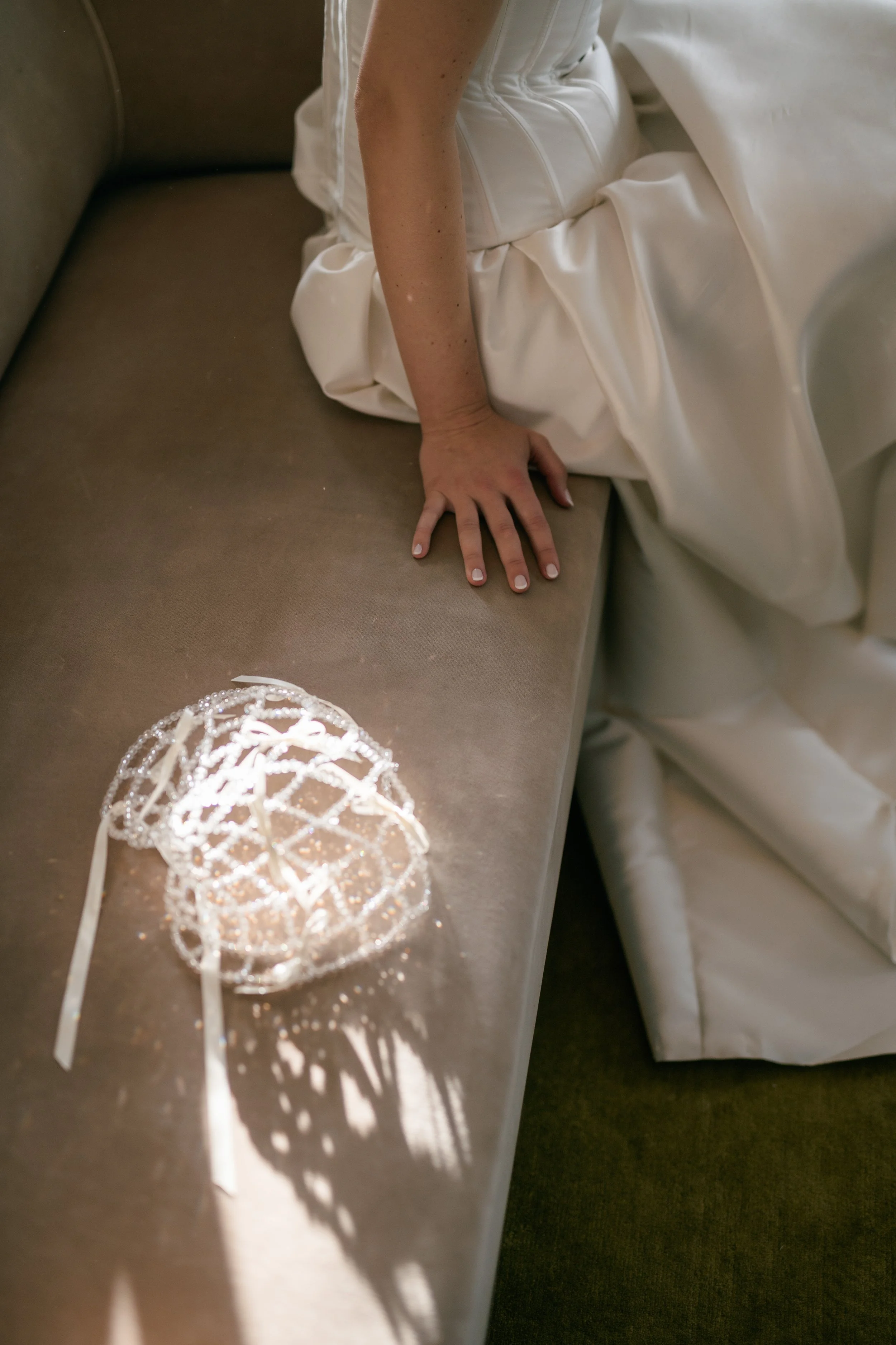 A bride in a white wedding dress sitting on a beige couch with her hand resting on the cushion. A decorative tiara with beads and ribbons is on the cushion, casting a shadow.