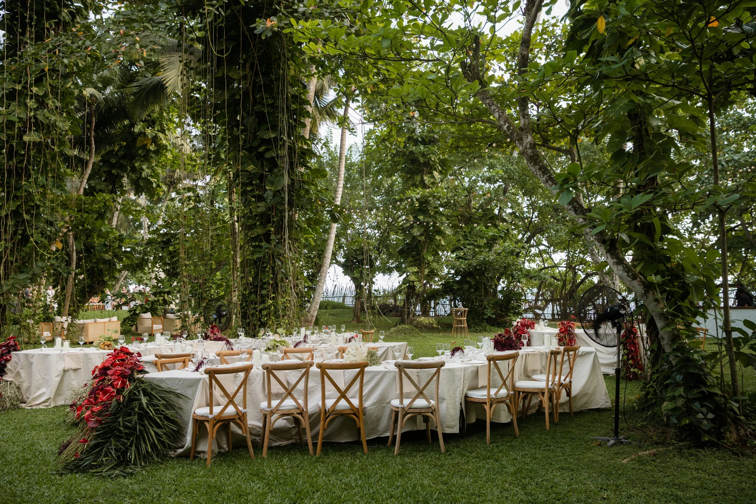 Outdoor banquet setup with banquet tables covered in white cloths, decorated with floral arrangements, surrounded by wooden chairs, amid lush green trees and foliage.
