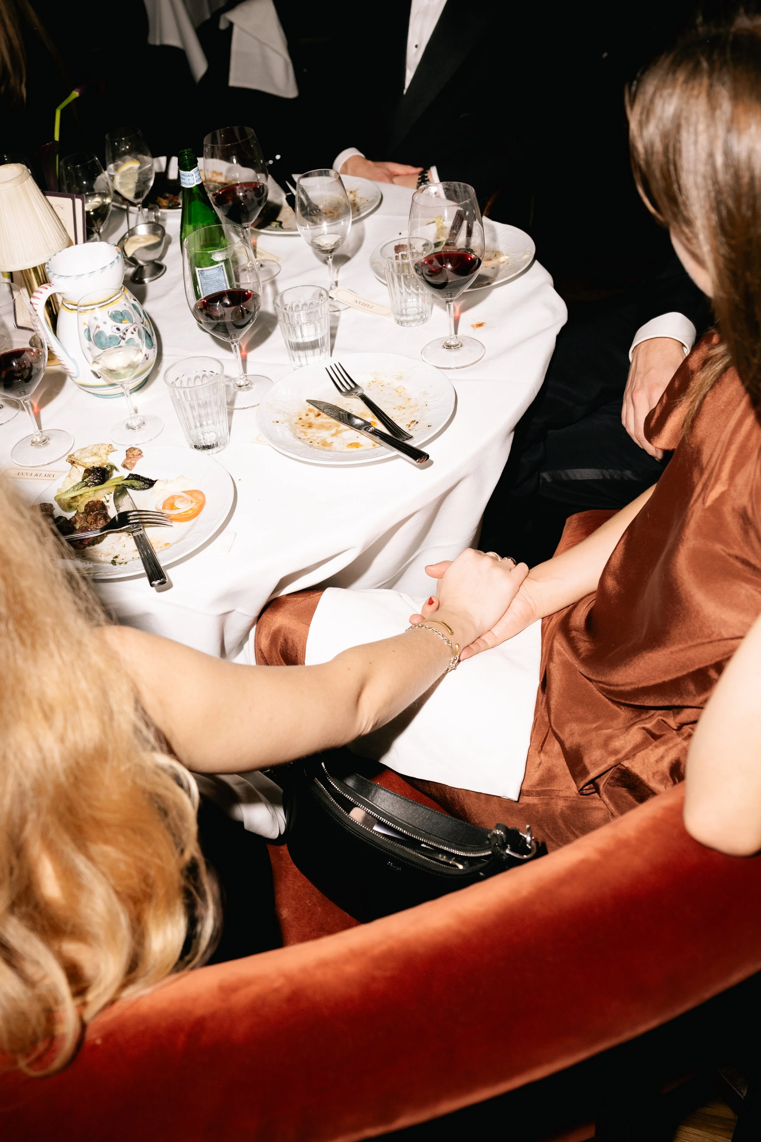 People holding hands at a dinner table with plates, wine glasses, and partially eaten food.