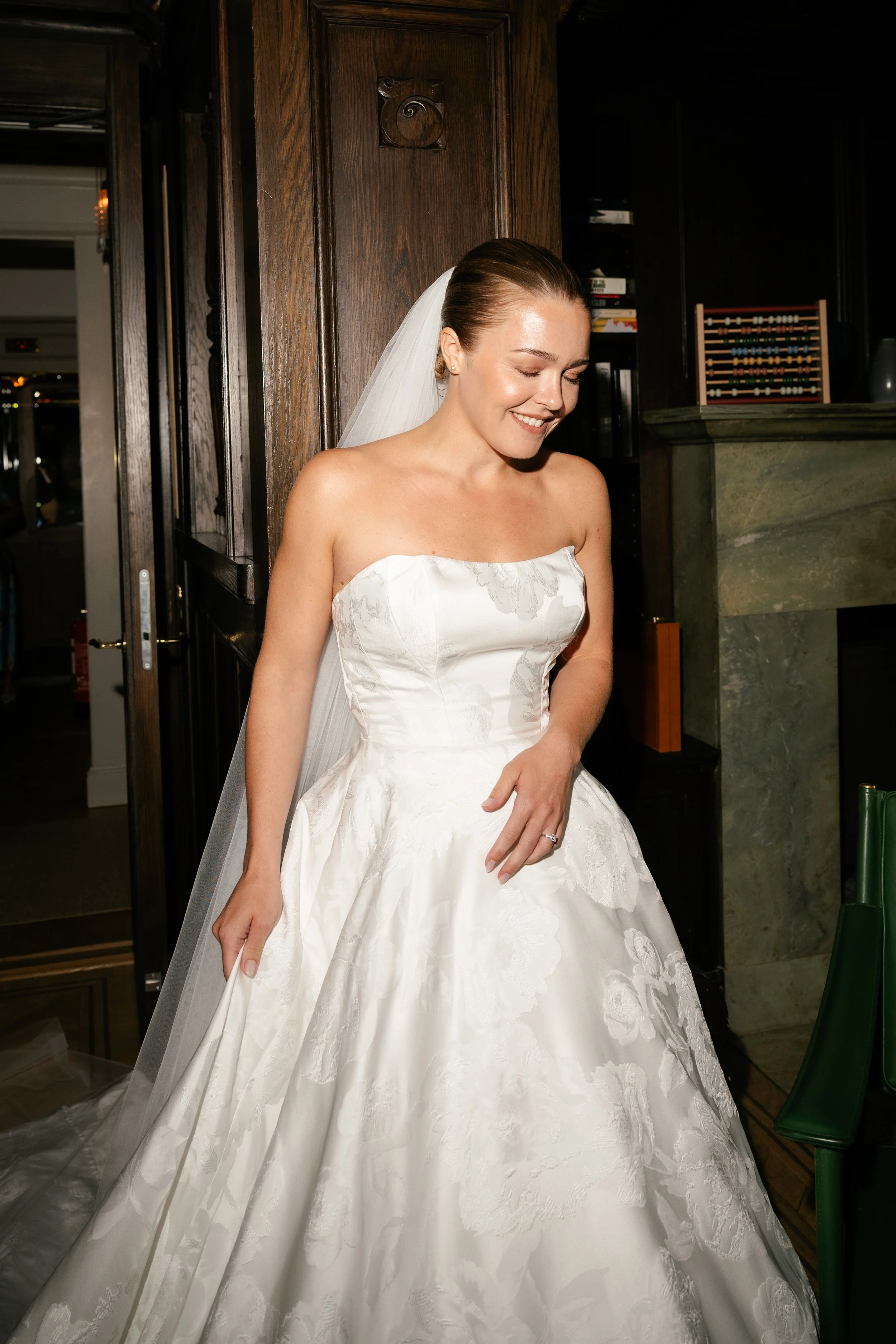 A bride in a strapless white wedding gown standing indoors, smiling with eyes closed, wearing a veil and a ring.