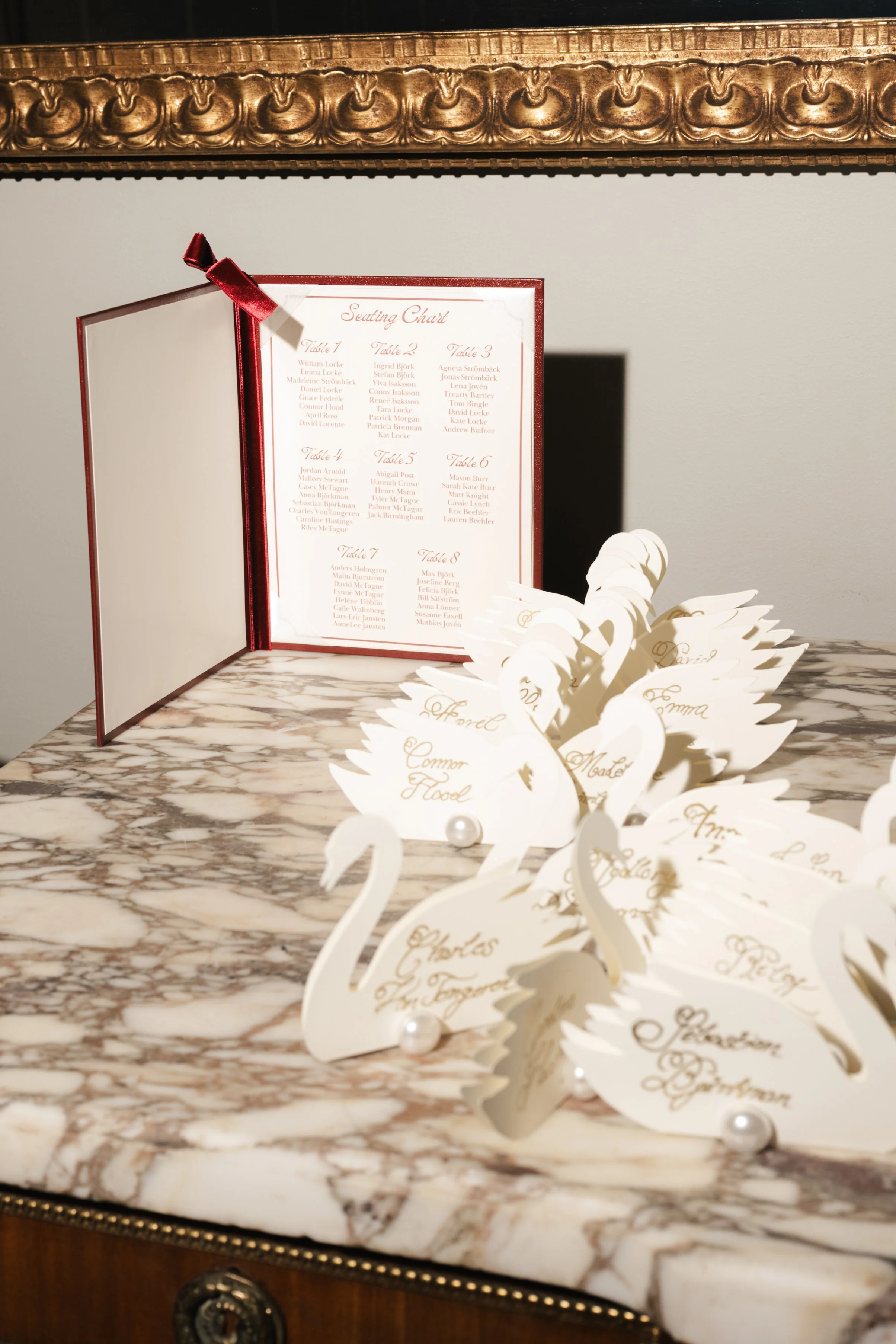A wedding seating chart with a red border and a red ribbon on the top left corner, placed on a marble table. Several white, paper swan-shaped place cards with handwritten names are arranged in front of the chart.