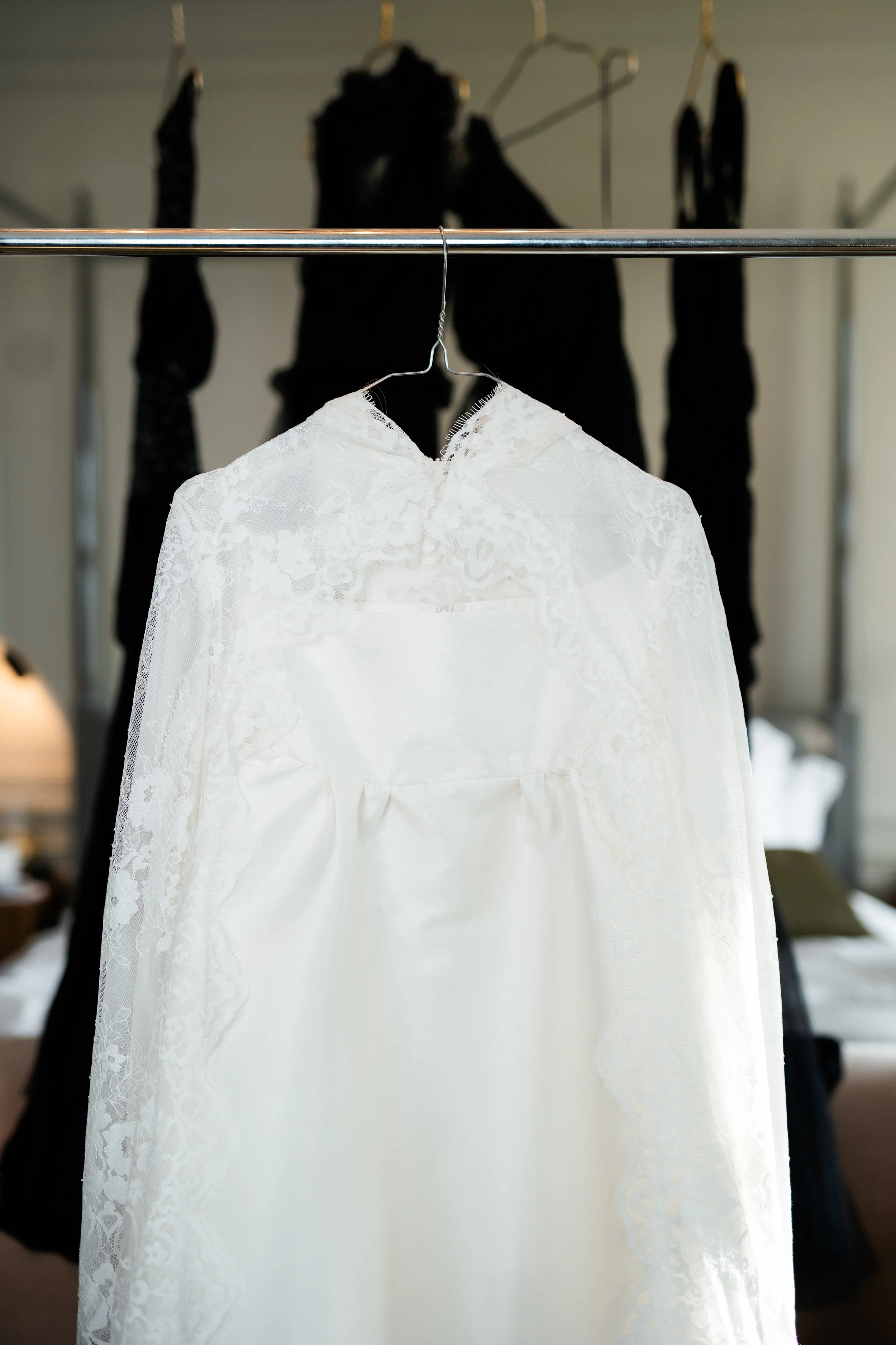 A white lace wedding dress hanging on a metal clothing rack with black clothing visible in the background.