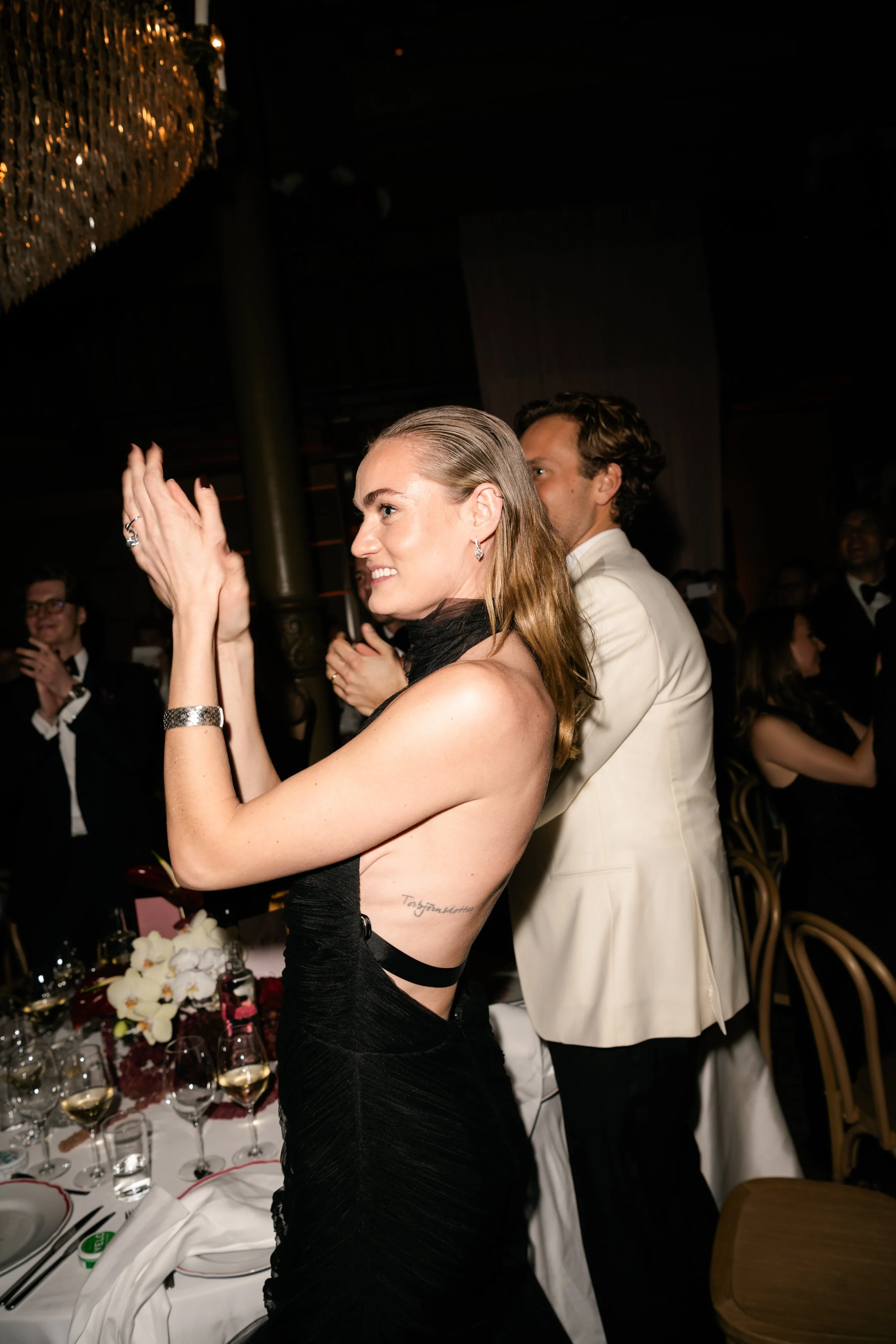 Woman in a black dress clapping at a formal event, with other guests in formal attire in the background.