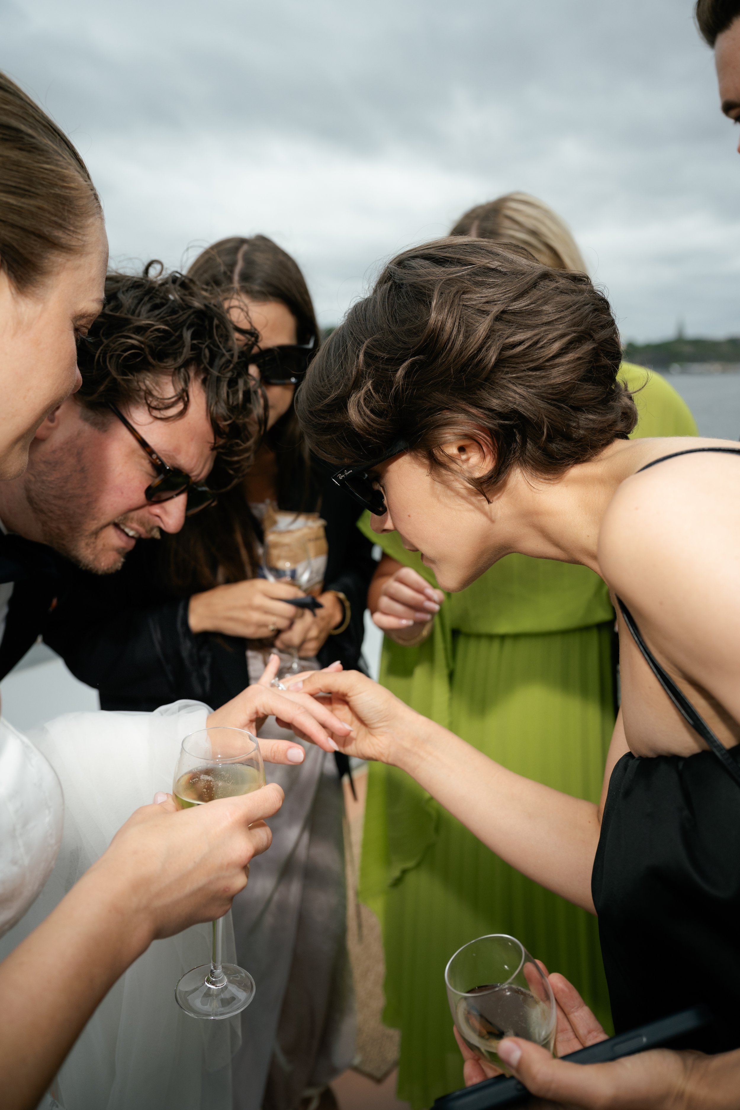 Group of friends socializing outdoors, some wearing sunglasses, holding glasses of champagne, with cloudy sky and water in the background.