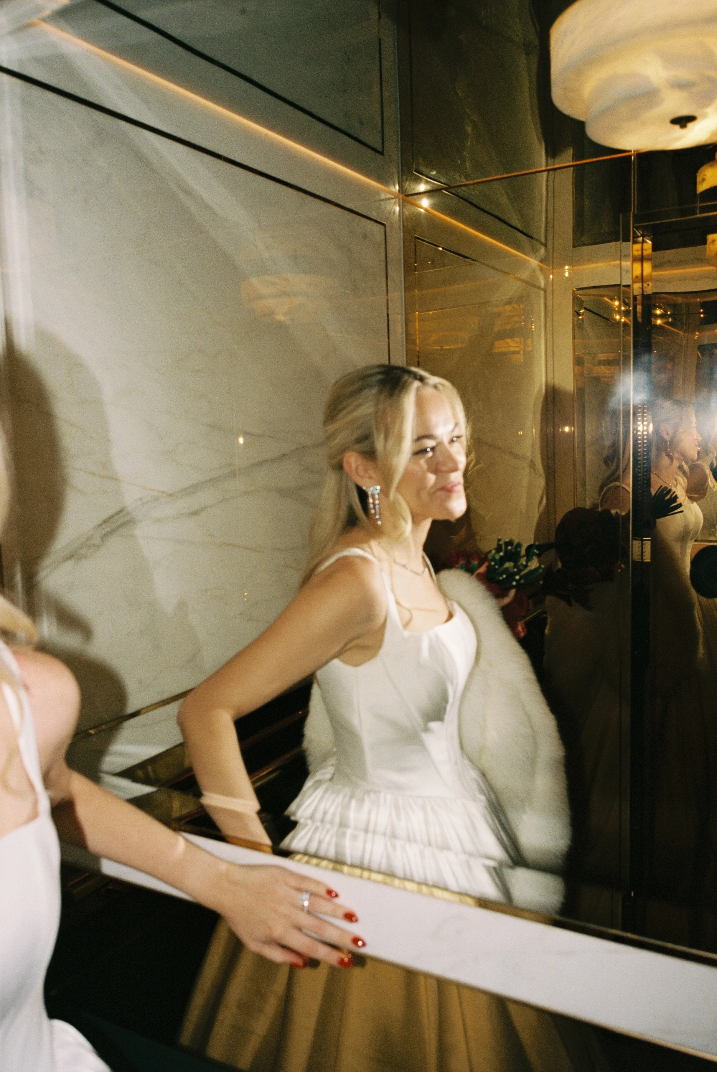 A woman in a white dress is standing in front of a mirror, smiling. She is wearing earrings and has blonde hair, with her reflection visible in the mirror.
