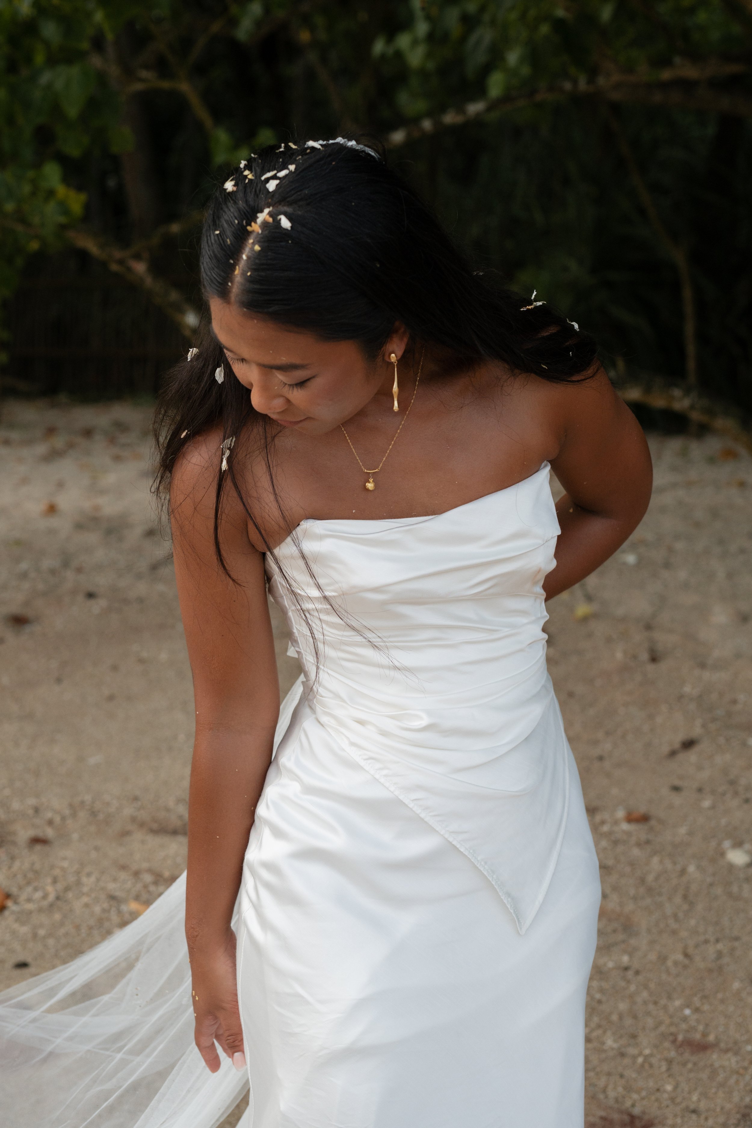A woman in a strapless white wedding dress, with long dark hair, is looking down with her hand on her hip, on a beach with trees in the background, with flower petals in her hair.