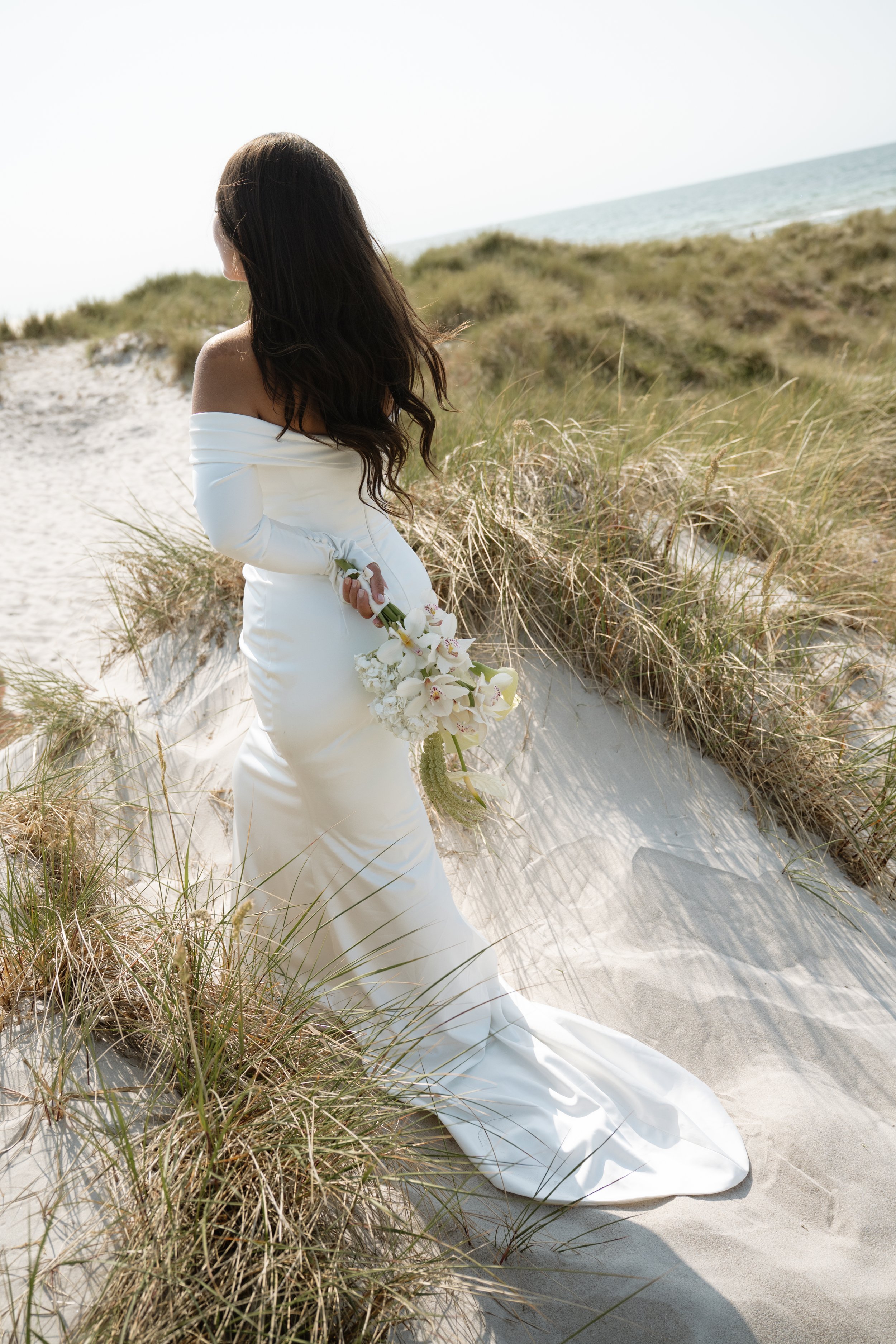 A woman in a white off-the-shoulder wedding dress standing on a sandy beach with grasses, holding a bouquet of white and pink flowers, facing away from the camera.