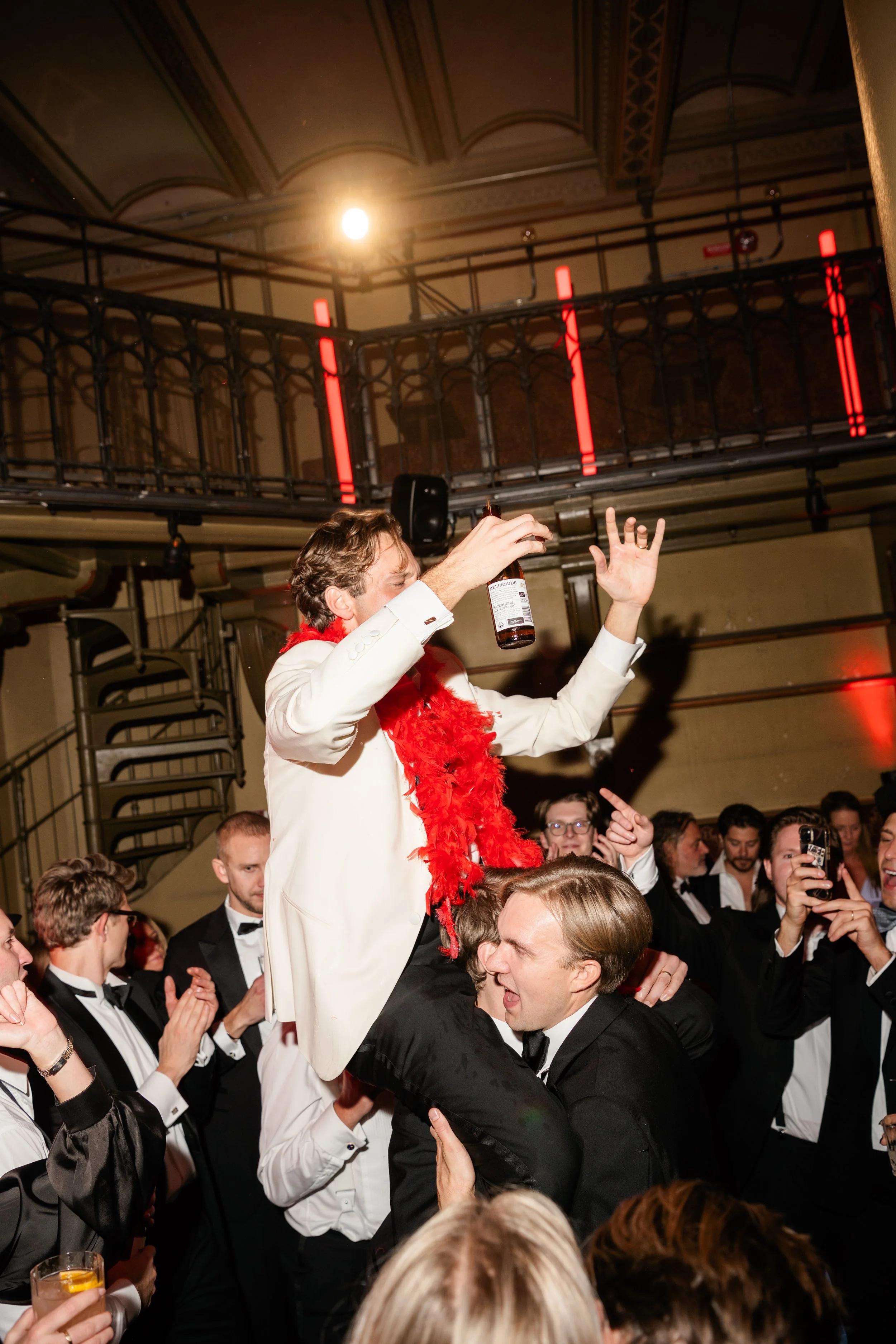 A man in a white suit with a red feather boa being lifted by another man during a celebration, surrounded by people in tuxedos at a party or event.